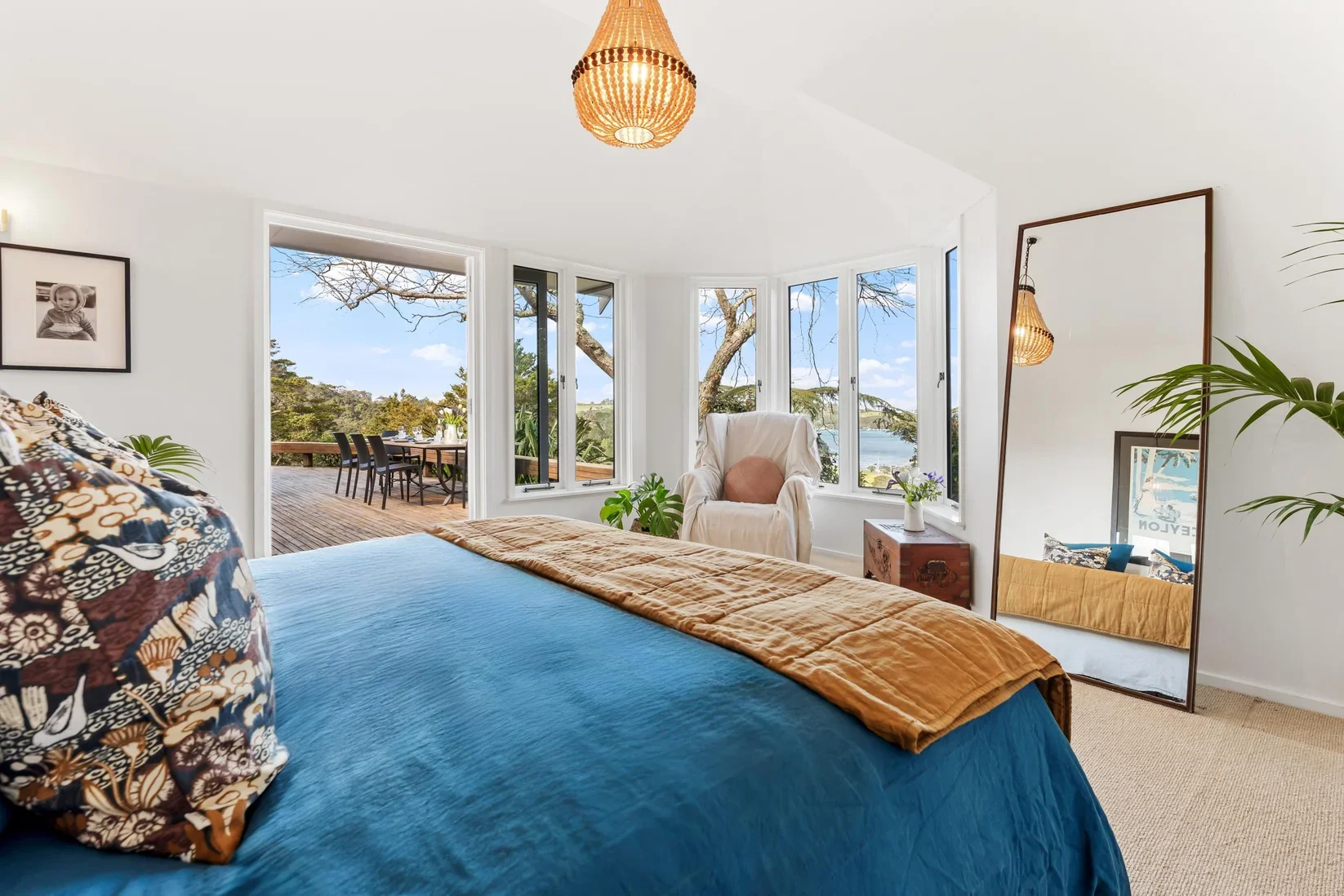 Bright bedroom with a large bed covered in blue and patterned pillows, overlooking a wooden deck with outdoor dining, and surrounded by windows revealing a view of trees, water, and a blue sky.
