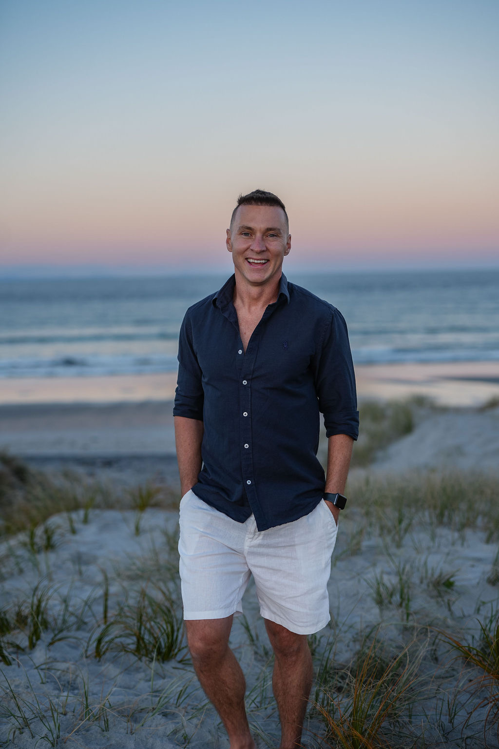 A man standing on a beach during sunset, wearing a navy blue shirt and white shorts, smiling at the camera.