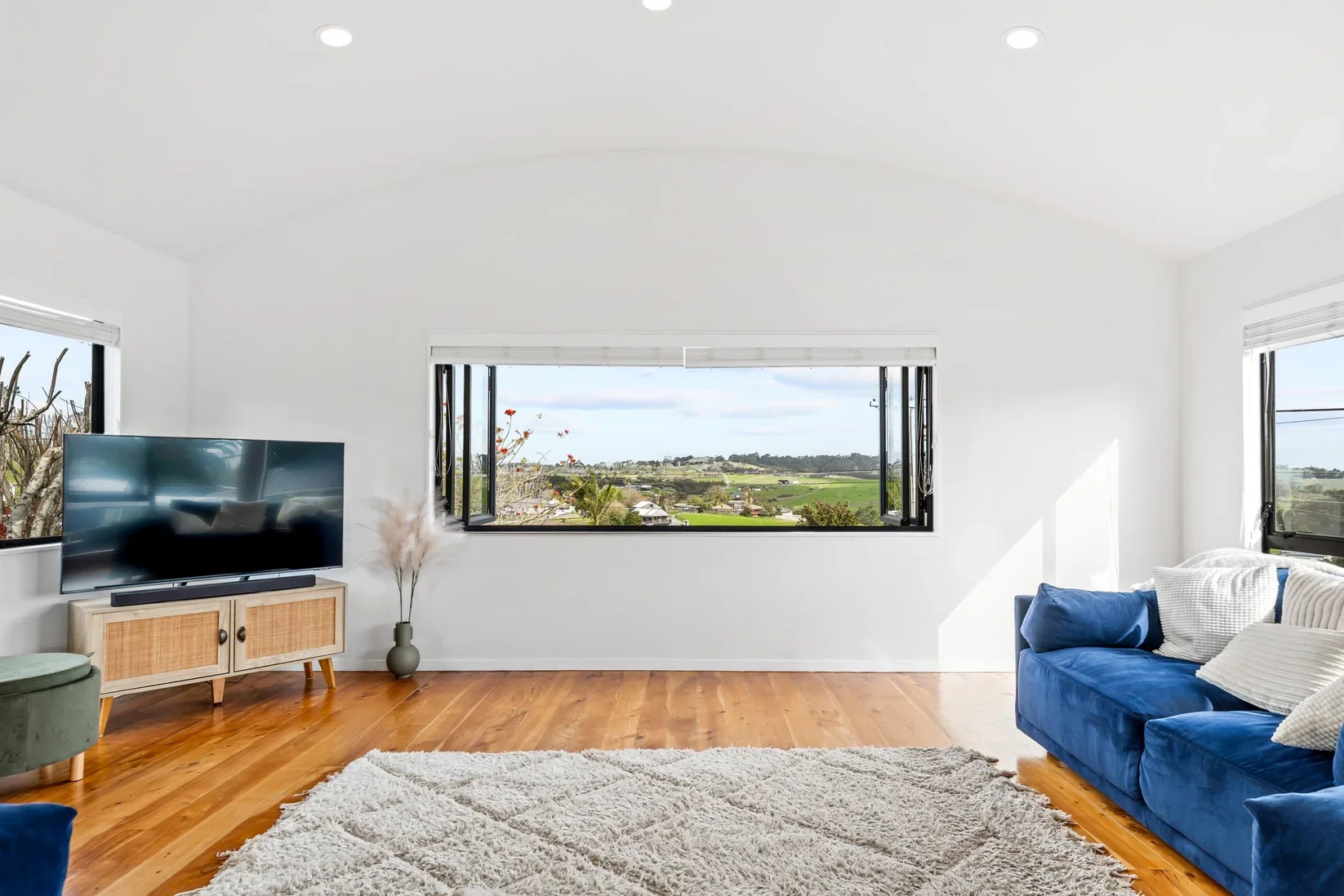 Living room in Snells Beach with white walls, wooden floor, a blue sofa with throw pillows, a television on a wooden stand, large windows showing a landscape view of fields and hills.