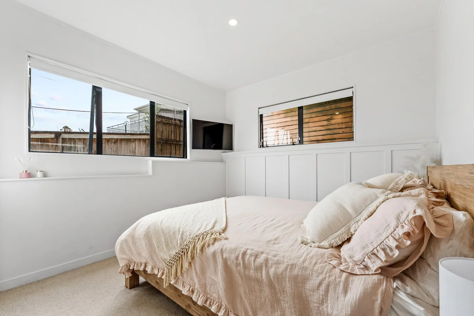 A minimalist bedroom with a neatly made bed covered in blush pink bedding, beige pillows, and a throw blanket. The room has white walls, a beige carpet, and two windows showing an outdoor wooden fence and neighboring buildings. A small flat-screen TV