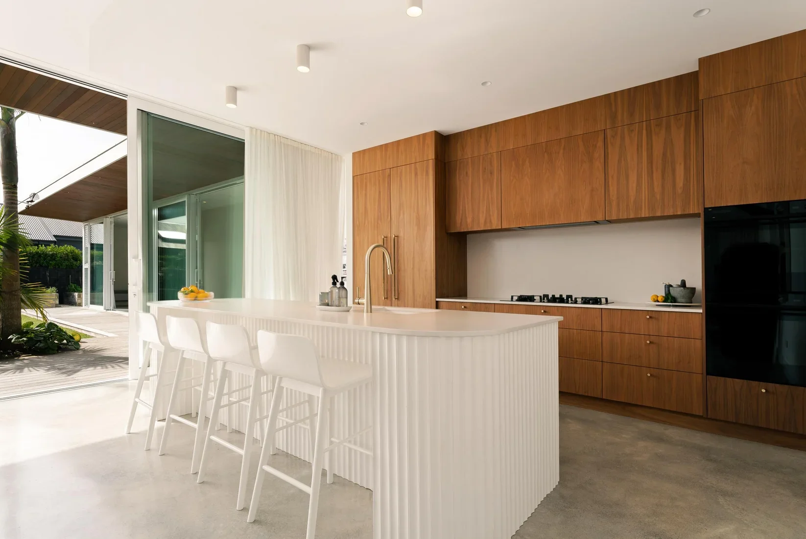 Modern kitchen with white island counter, four white bar stools, wooden cabinets, built-in stove, and large sliding glass door with curtains that open to outdoor patio and greenery.