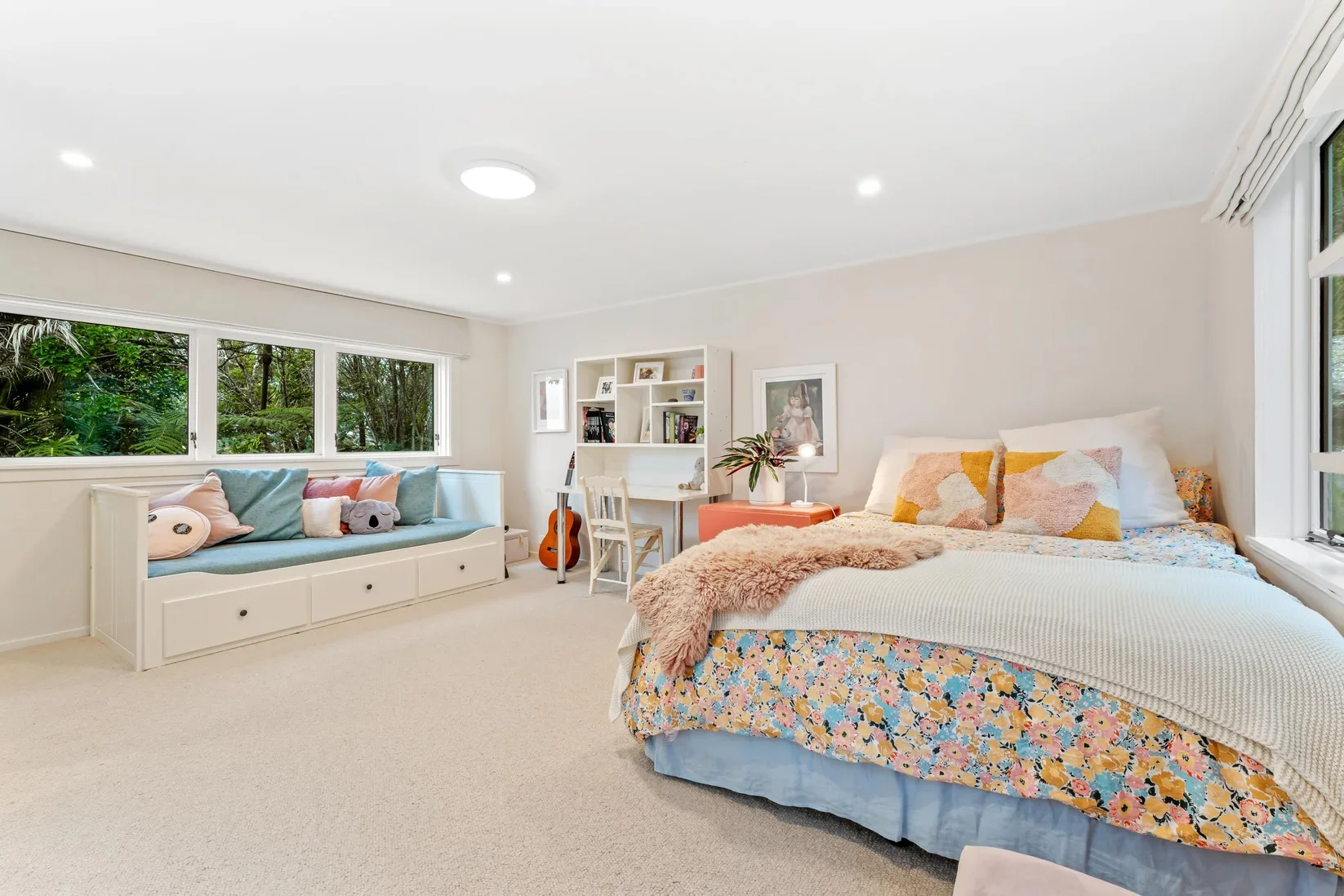 Bright bedroom with a bed covered in a floral quilt, pillows, and a plush throw, a window seat with soft cushions, a guitar, a white bookshelf, and decorative plants and artwork.