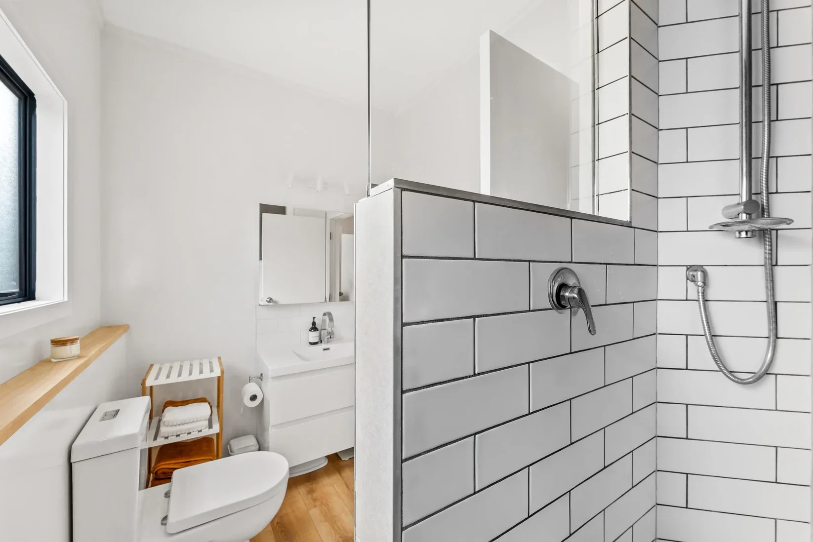 Modern bathroom with white walls, a window, a toilet, a small white sink with a mirror, and a separate shower area with white subway tiles and a chrome showerhead.