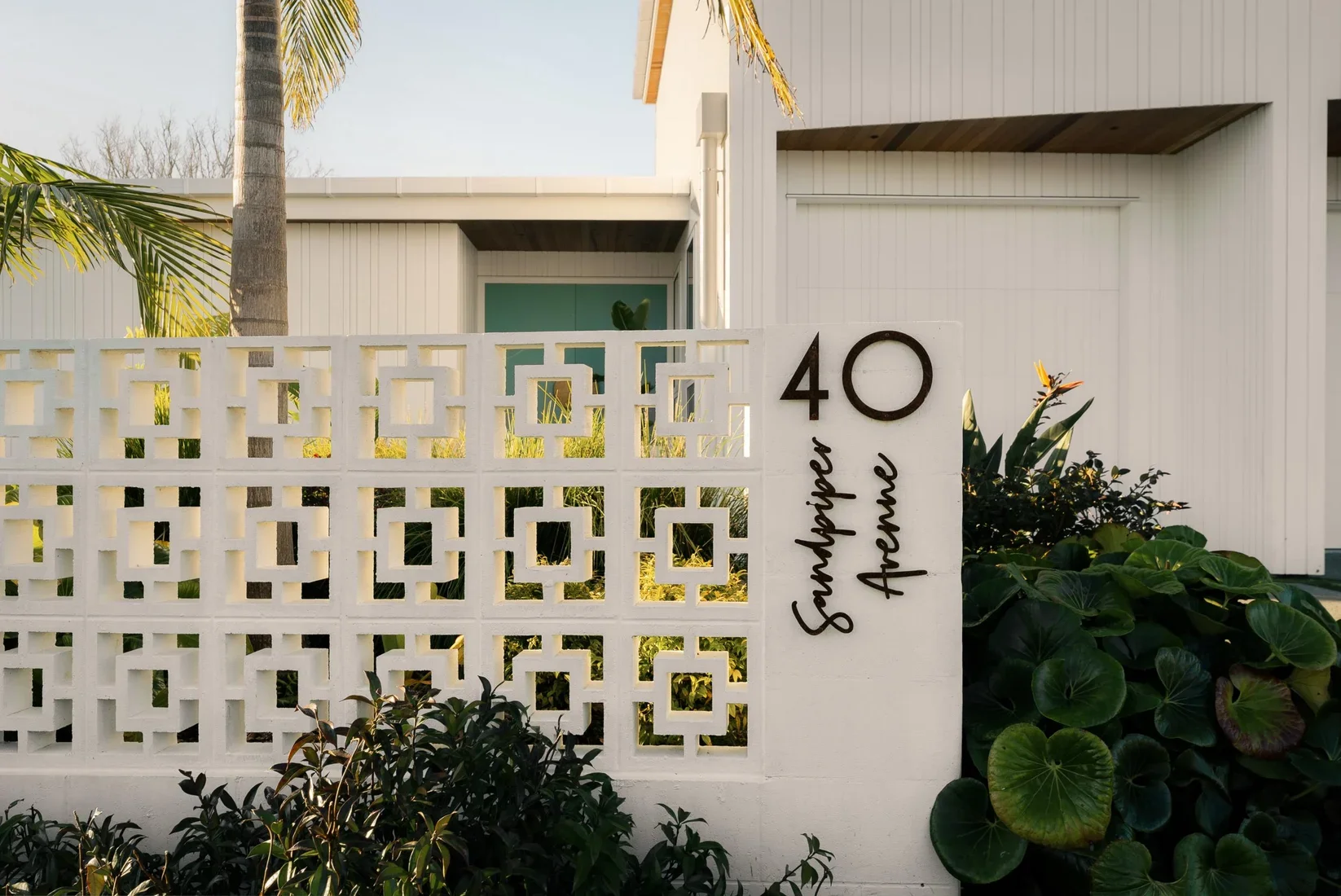 A white decorative wall with geometric cutouts in front of a modern house. The house has white paneling and an overhang, with a palm tree nearby. There are plants with round leaves at the base of the wall. The house number "40" and the words "Sunshower Avenue" are on a white column next to the wall.