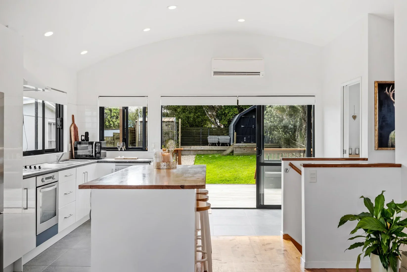 Bright modern kitchen with white cabinets, wooden countertop island, black-framed windows, sliding glass door leading to a green backyard, and indoor plants.