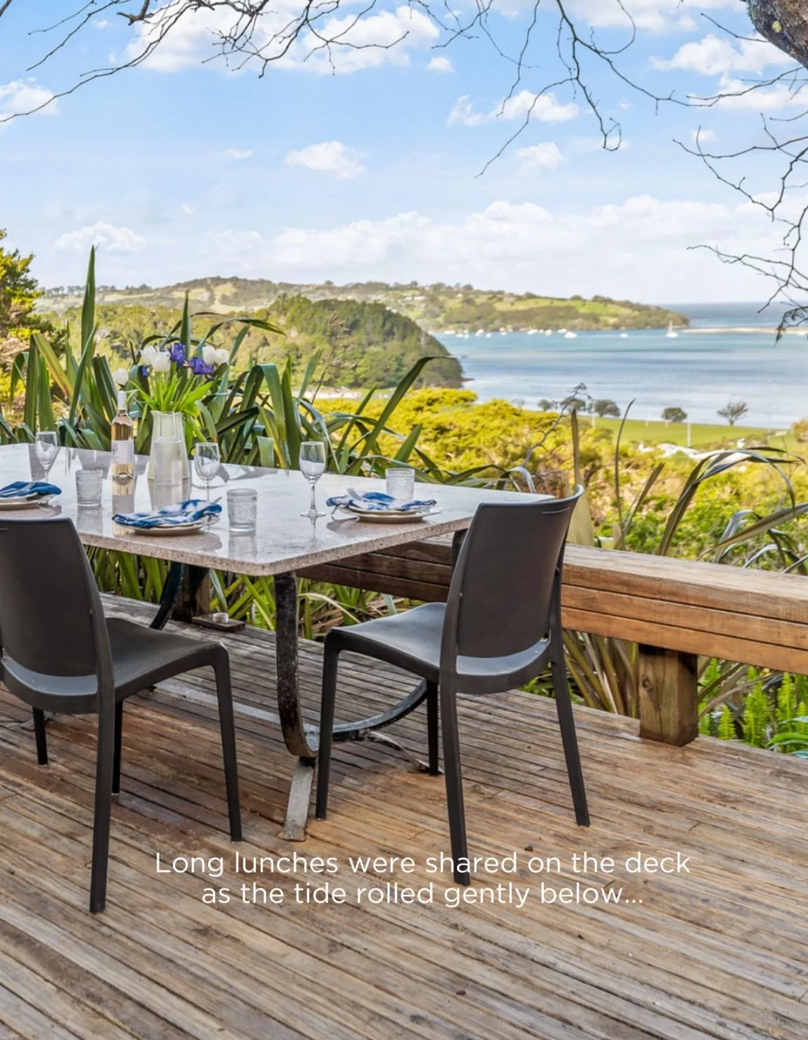 Outdoor dining table set on a wooden deck overlooking a scenic coastline with green hills, water, and boats, under a cloudy sky.  596 Leigh Road, Whangateau.