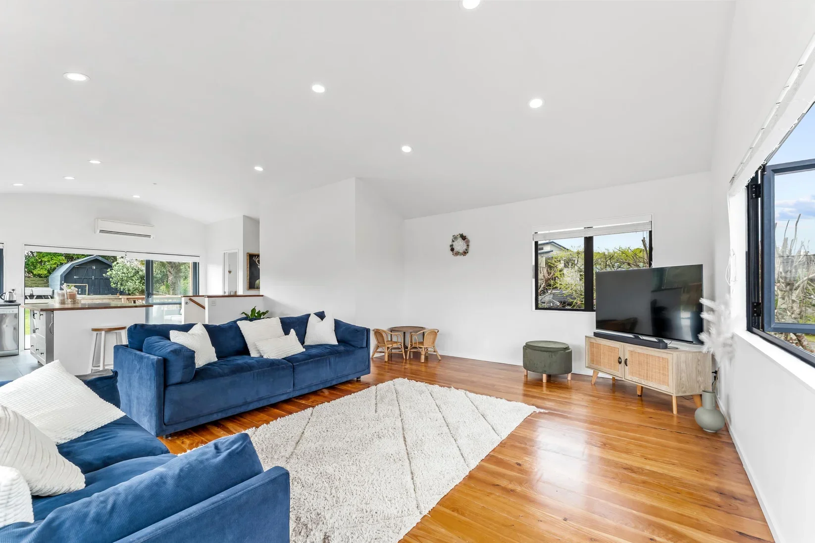 A home in Snells Beach with bright living room with white walls, hardwood floors, and large windows. Contains blue velvet sofas with white pillows, a white area rug, a TV on a wooden stand, and a small green ottoman.