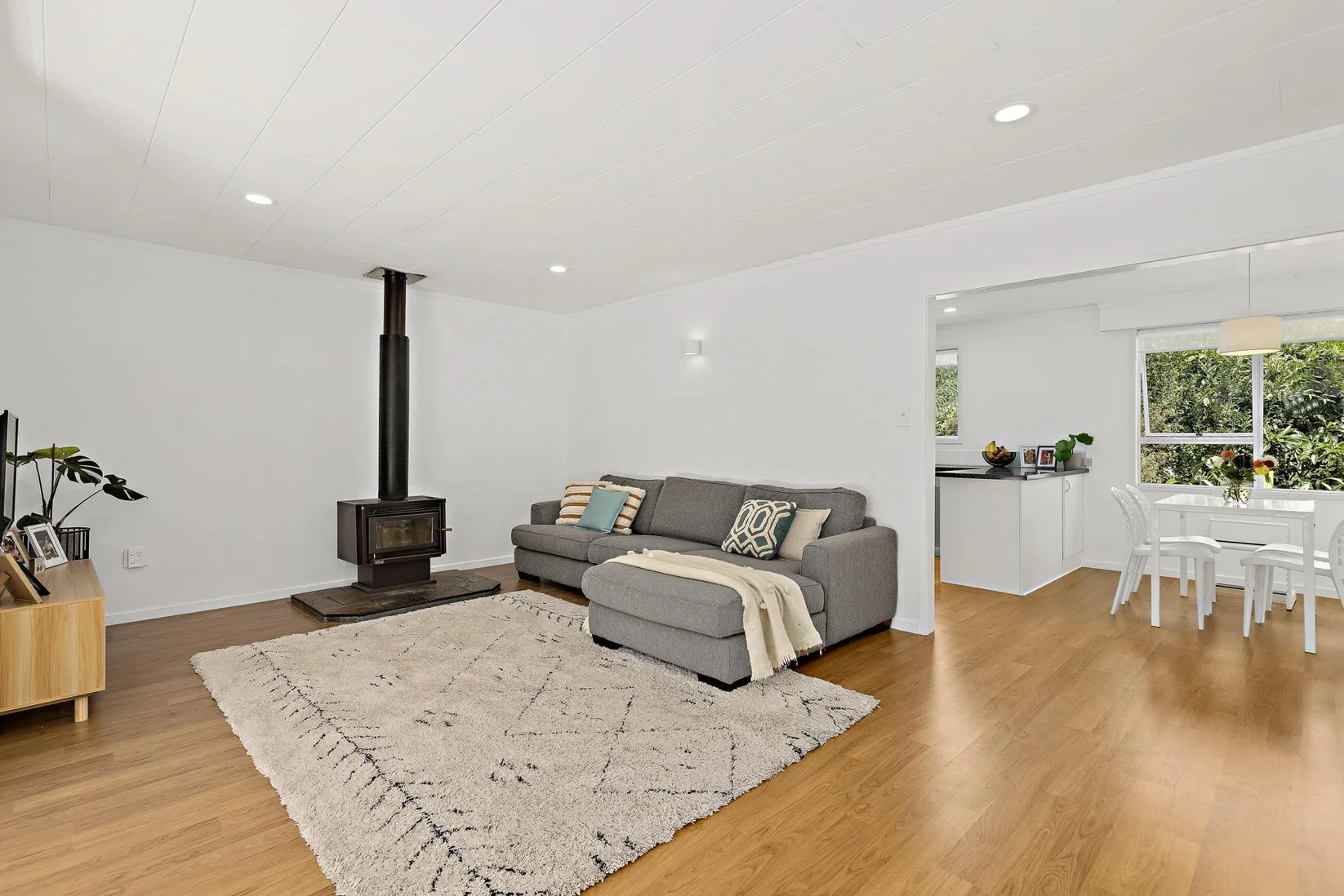 Modern living room with gray sofa, cream and patterned pillows, beige rug, white walls, wood flooring, and wood-burning stove