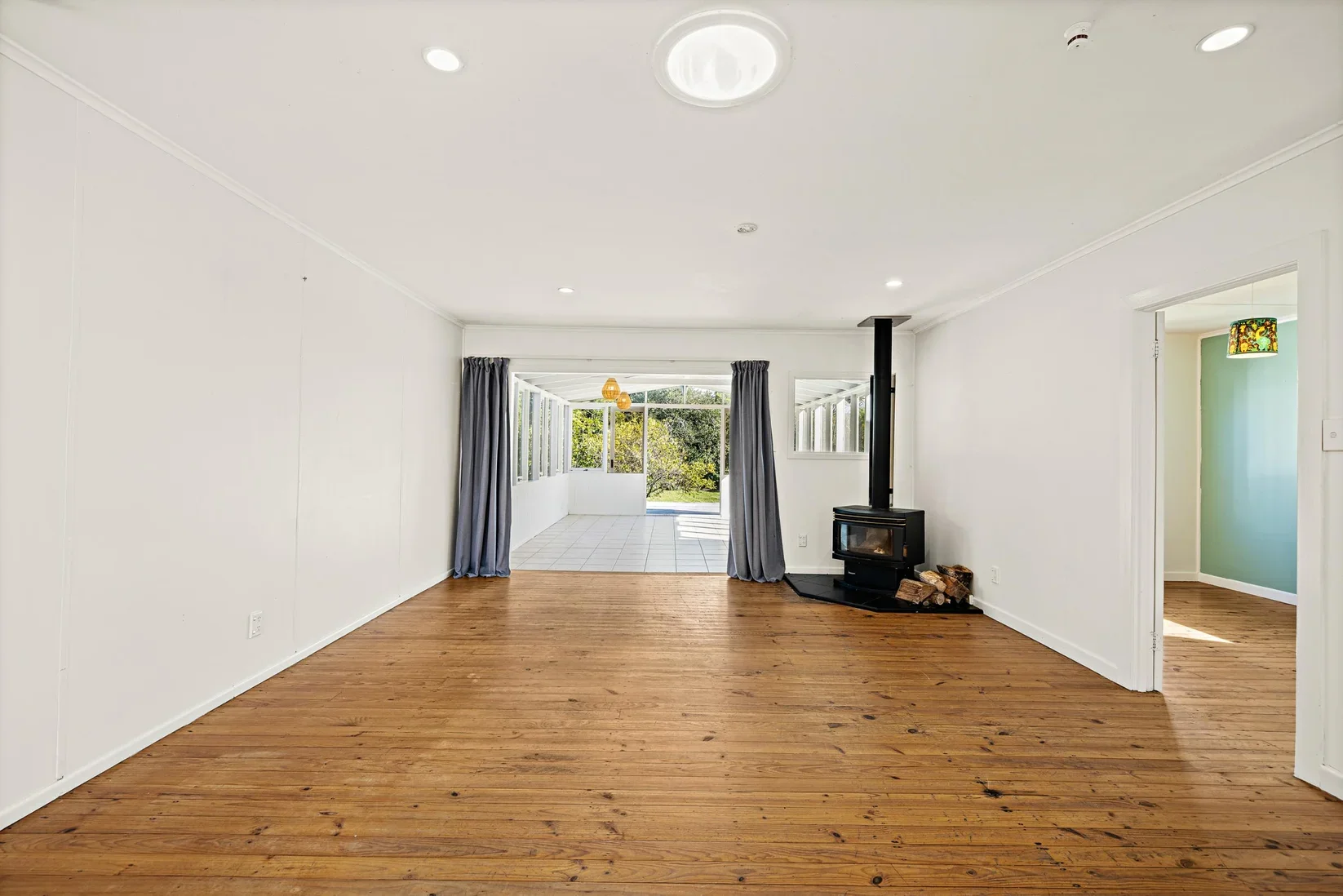 Empty living room in Point Wells home with hardwood floors, white walls, a black wood stove, and a view of a patio with large windows and sliding glass door.