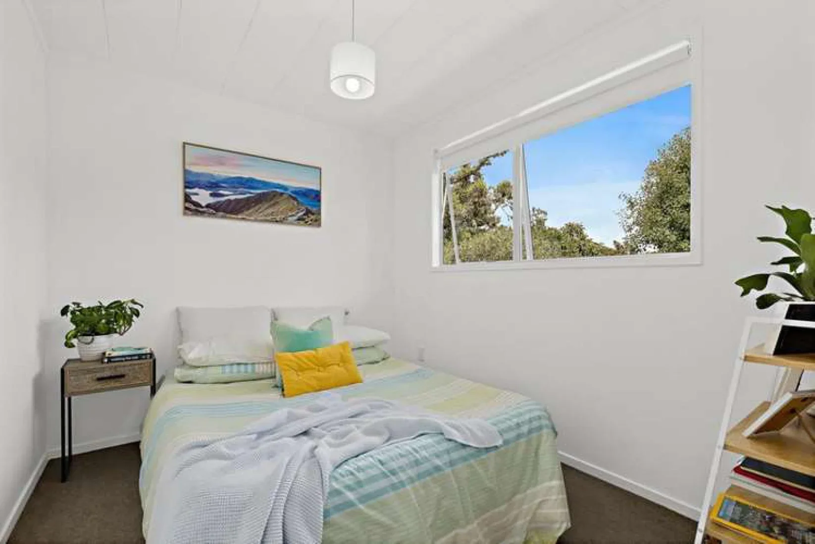 A small bedroom with a bed covered in light-colored striped bedding, two pillows, and a blanket. There's a nightstand with a potted plant and books on it, and a white shelving unit with books, a laptop, and a plant. A window shows a view of trees and