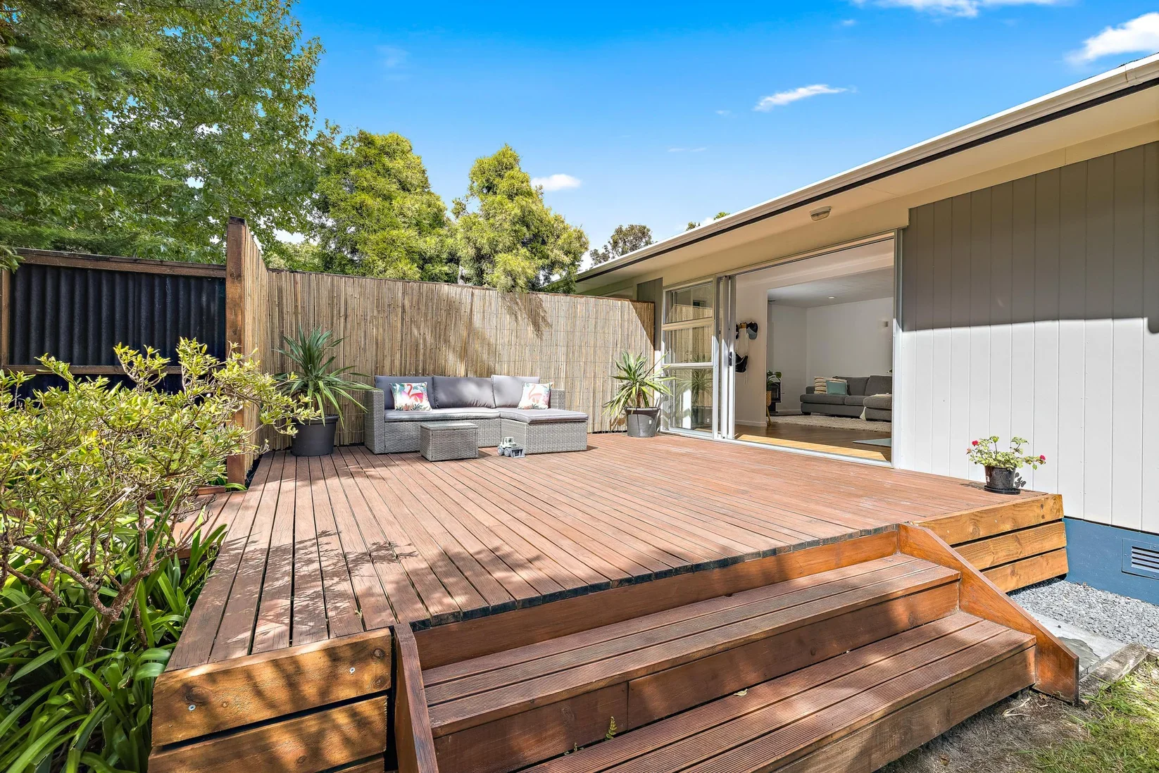 Backyard wooden deck with potted plants, outdoor seating, and open sliding glass door leading to living room.
