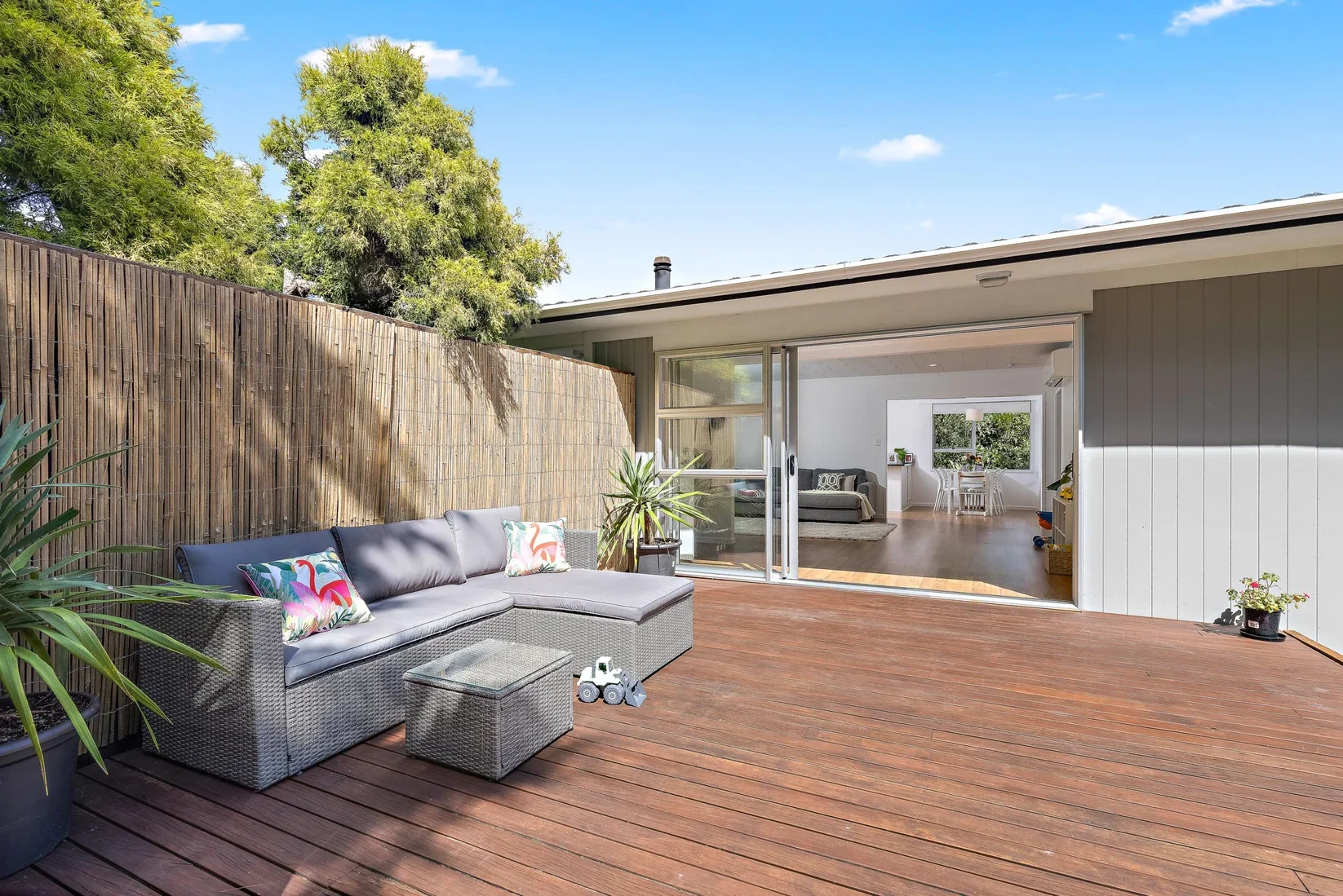 Outdoor wooden deck with a grey sectional sofa, colorful pillows, and a small glass-topped table. A toy truck is on the deck. The area is partially enclosed by a wooden fence, with trees in the background. The deck is attached to a house with sliding