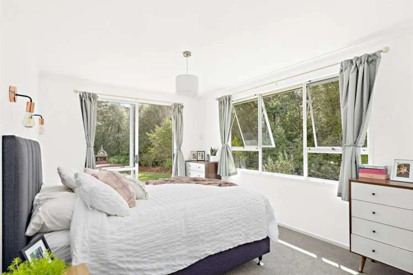 Bright bedroom with large windows, white walls, bed with gray upholstered headboard, white bedding, and several pillows, dresser with photo frame, potted plant, and pink books, outdoor view of trees and greenery.