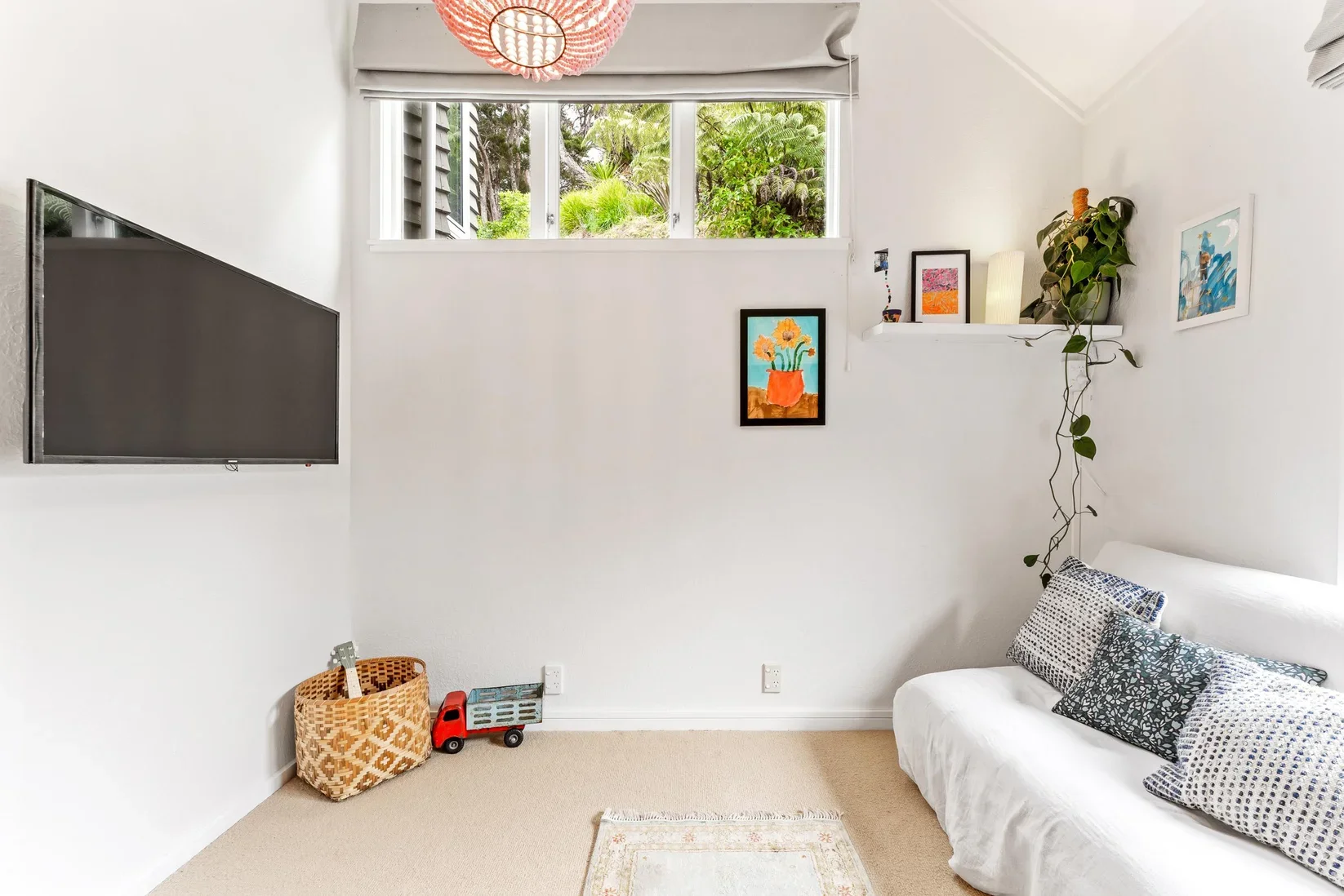 A bright white living room with a wall-mounted flat-screen TV, a small wicker basket with a toy and a toy truck on the floor, and a white sofa with patterned pillows near the corner. There is a small rug on the beige carpet. The wall has a high windo