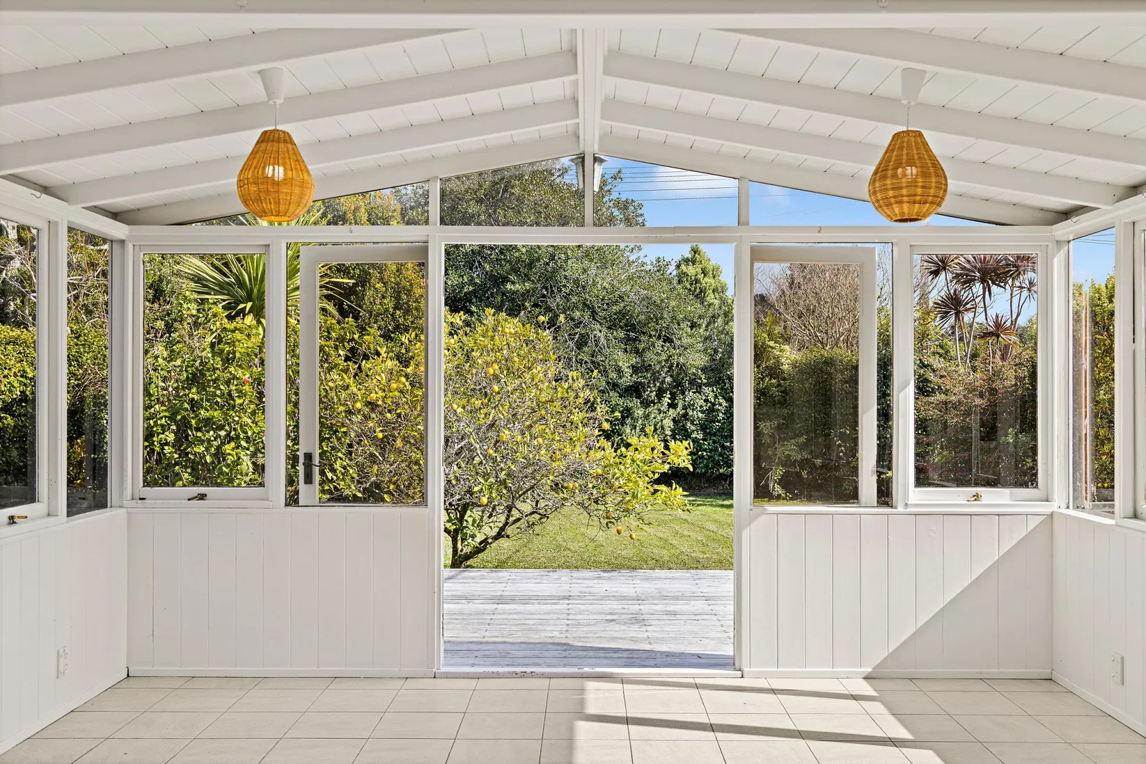 Sunlit enclosed porch with white walls and ceiling, hanging wicker light fixtures, and a view of a garden with trees and a lemon tree outside.
