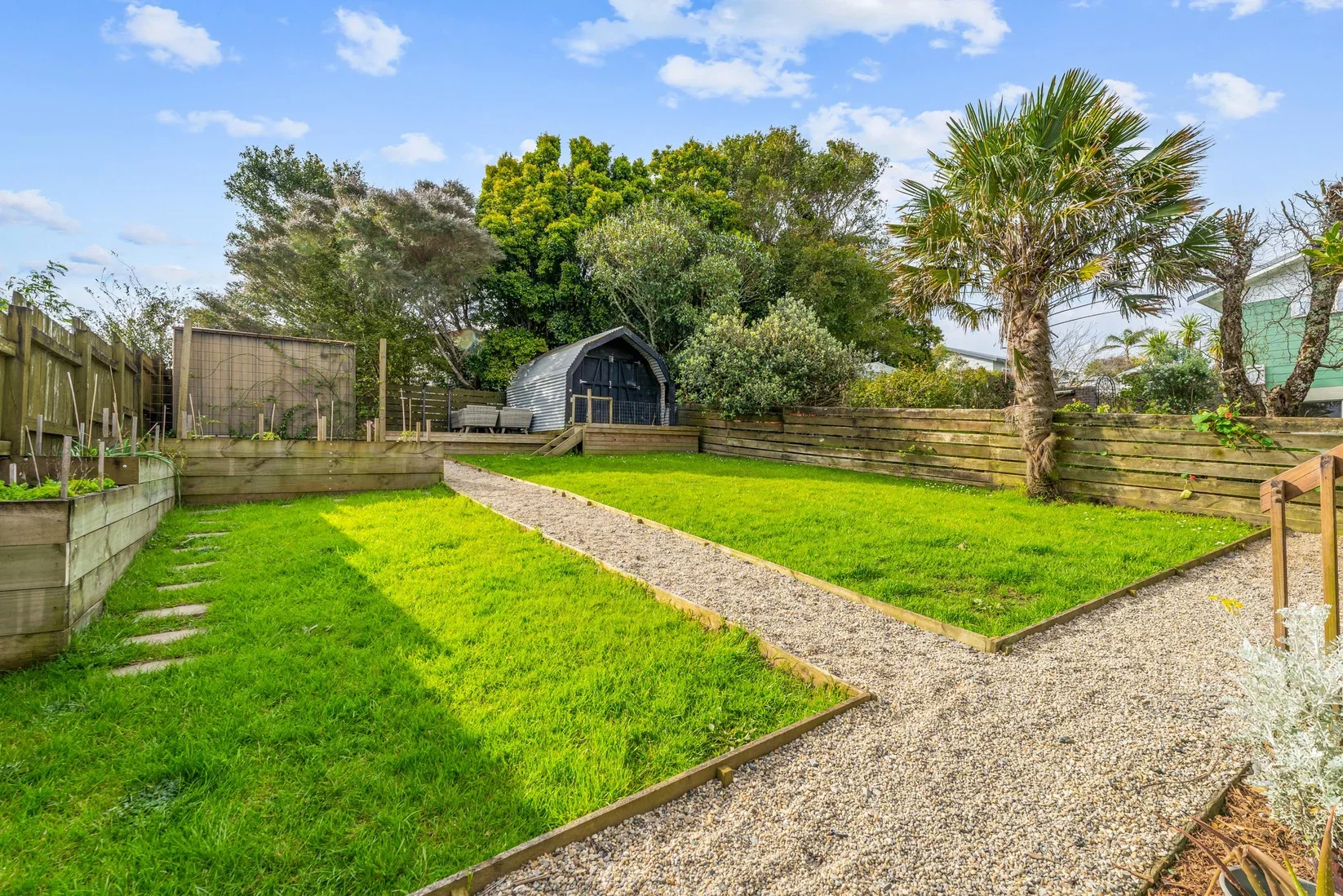 A backyard with a gravel pathway, green grass, raised garden beds, and a shed surrounded by trees and a wooden fence under a partly cloudy sky.