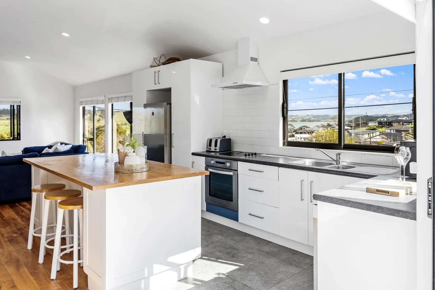 Modern white kitchen with black countertops, a window with a view of houses and water, a wooden dining table, and bar stools