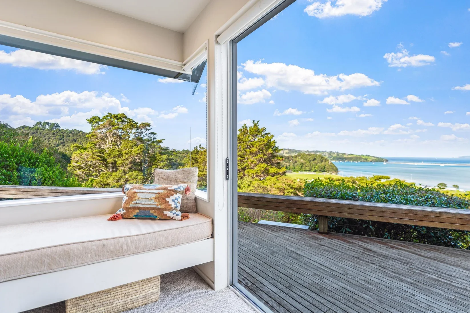 A living room nook with a cushioned window seat and decorative pillows, overlooking an outdoor wooden deck and a scenic view of trees, water, and blue sky.