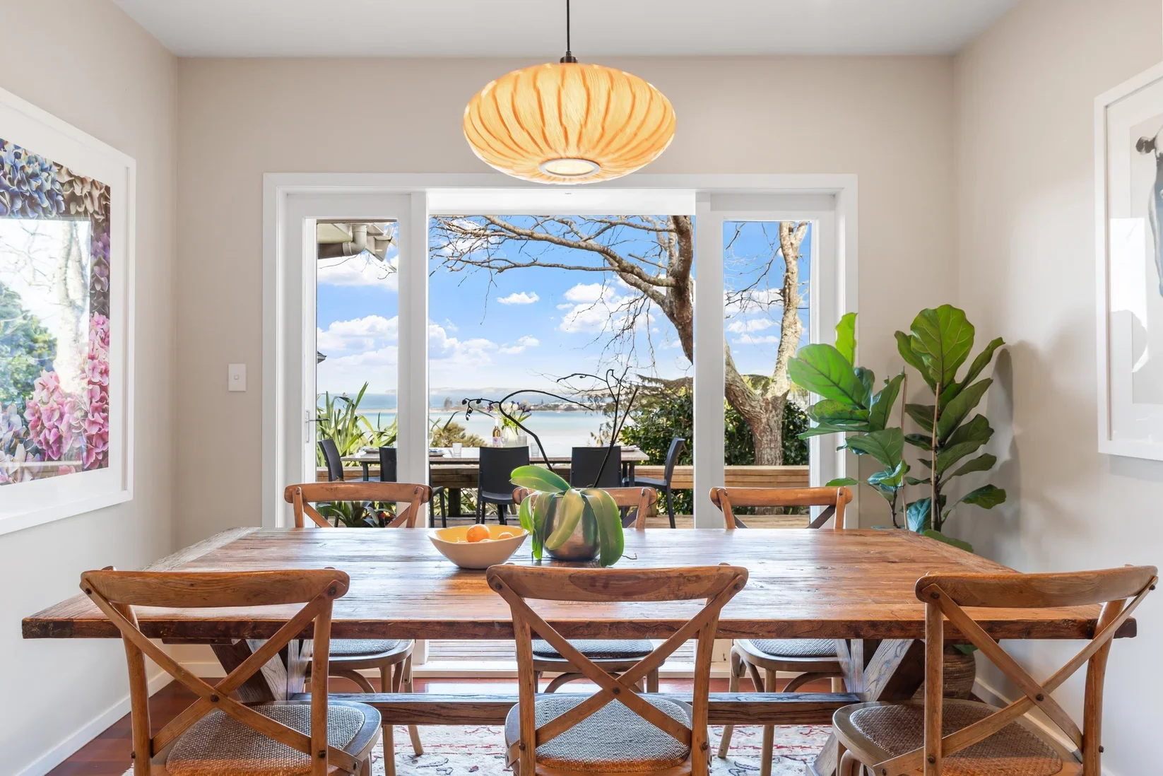 Dining room with a rustic wooden table, six wooden chairs, a bowl with fruit, and a potted plant. Large sliding glass doors lead to a deck outside with an ocean view, trees, and blue sky with clouds.
