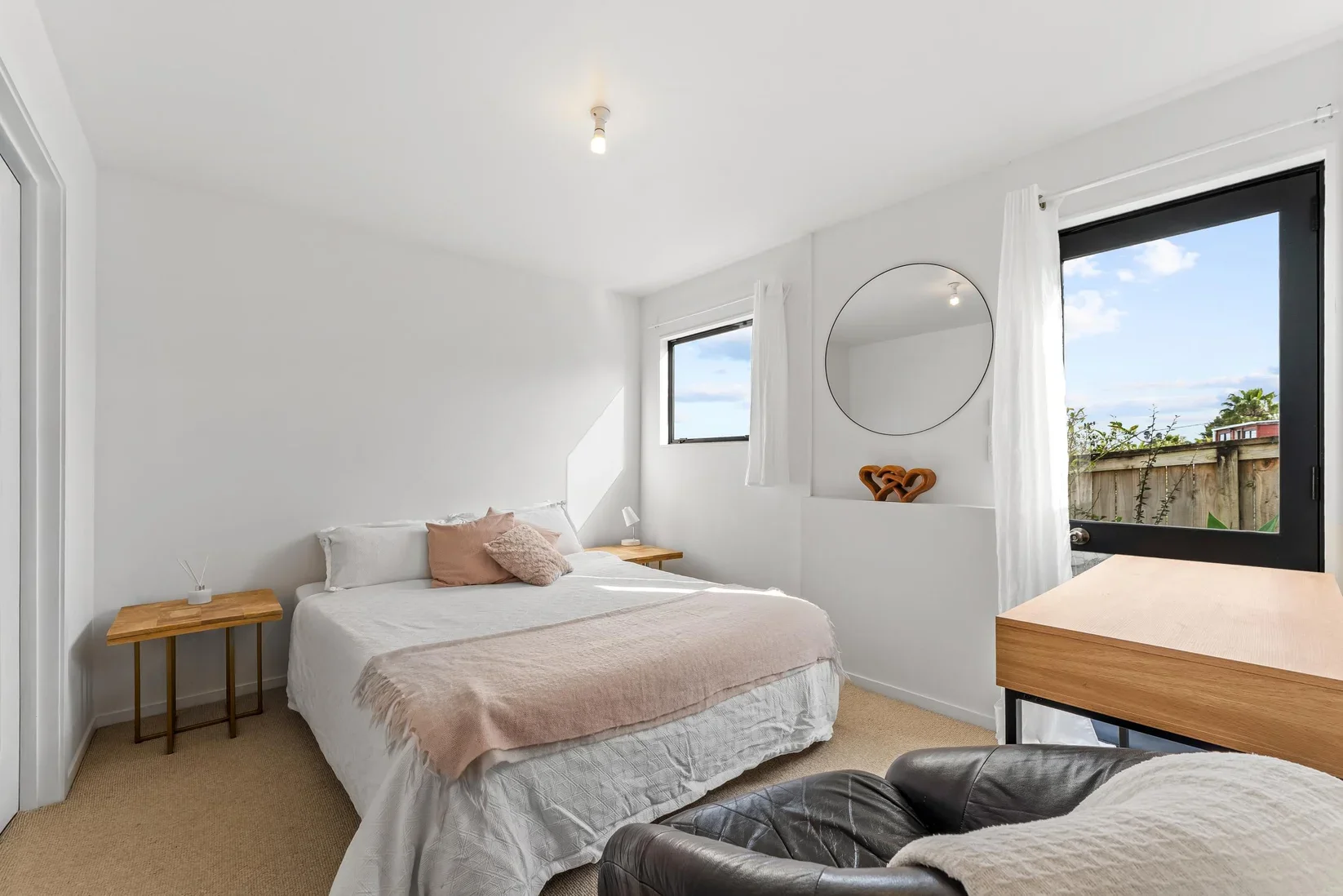 A bright, minimalist bedroom with white walls, a bed with white bedding and pink pillows, two wooden nightstands, a mirror on the wall, a window and a glass door with white curtains, a black leather chair, and a beige carpet.