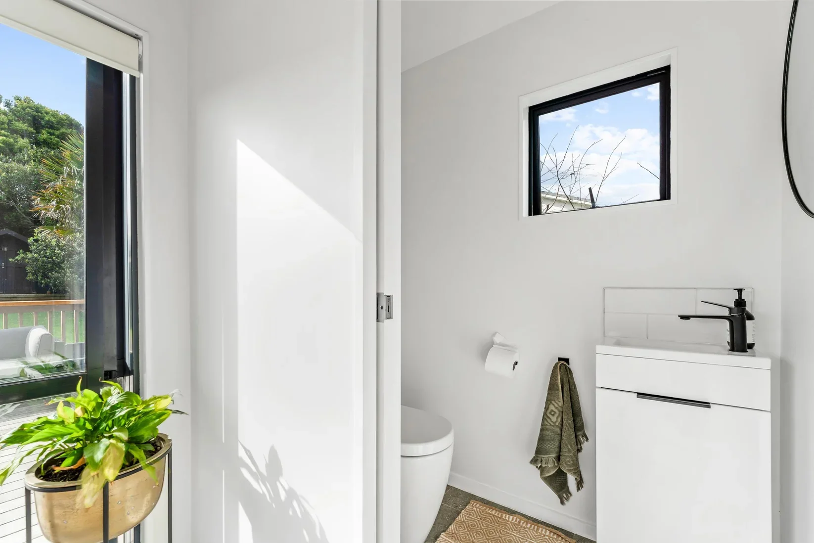 Small modern bathroom with white walls, a window showing trees outside, a white vanity with a black faucet, a toilet paper roll, a towel hanging, a mat on the floor, and a potted plant near a sliding glass door.
