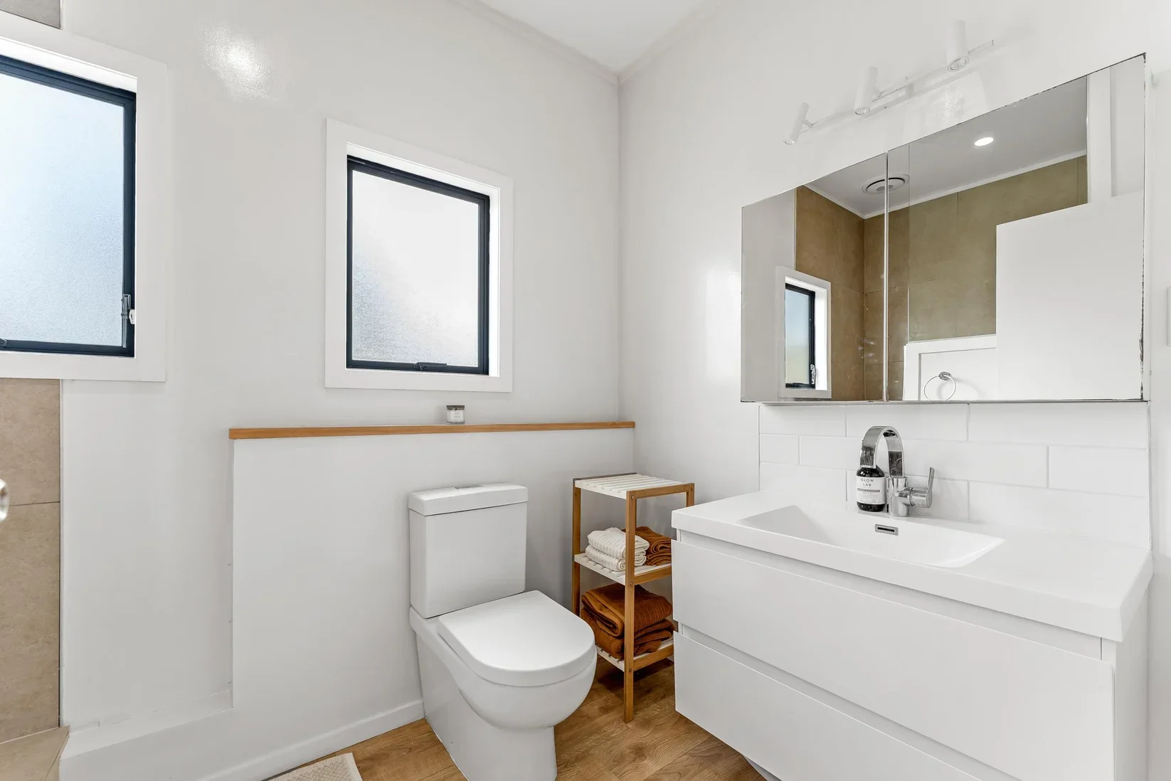 Modern bathroom with white walls, two frosted windows, a white toilet, a wooden shelf with towels, a white sink and mirror, and a large wall mirror.