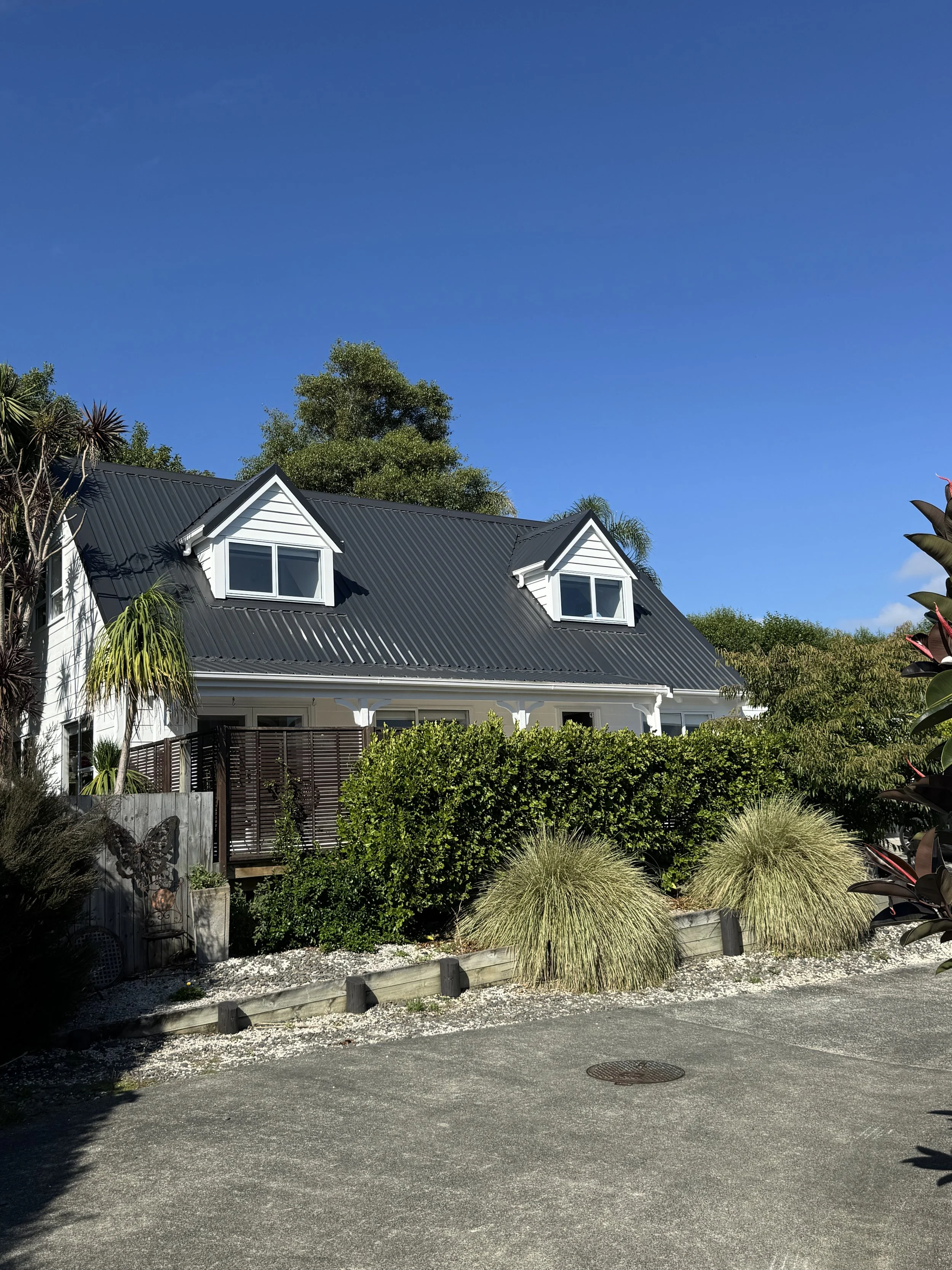 A two-story house with a black metal roof, white siding, and dormer windows. Surrounded by lush green shrubs and plants under a clear blue sky.
