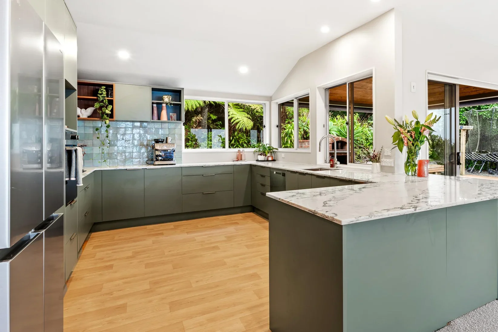 Modern kitchen with green cabinetry, marble countertops, wood flooring, and large windows overlooking greenery.