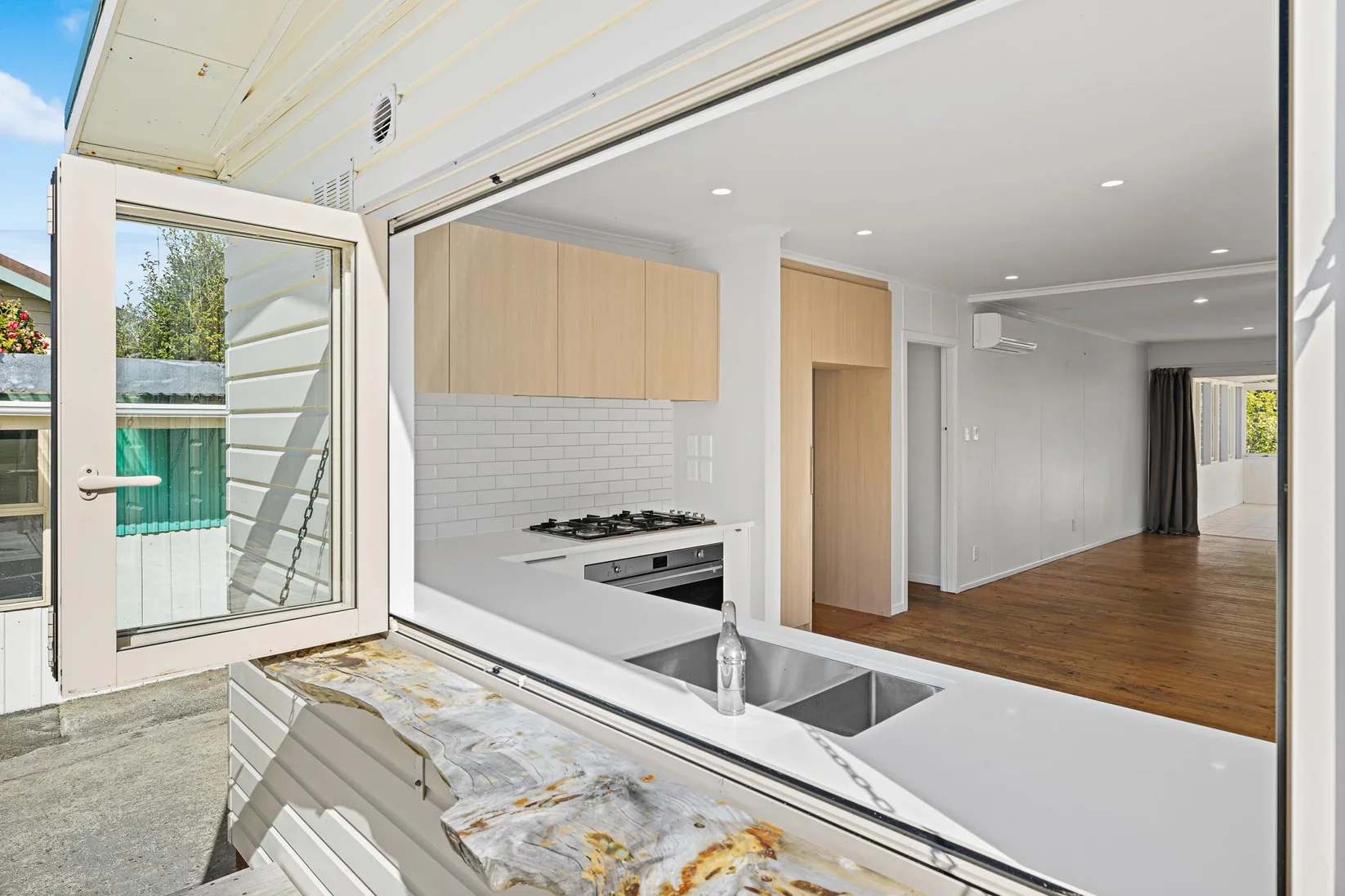 View of a modern open-concept kitchen in Point Wells from an outdoor patio, showcasing a white countertop with sink and faucet, light wood cabinets, white tiled backsplash, and visible living space with hardwood floors and large window.