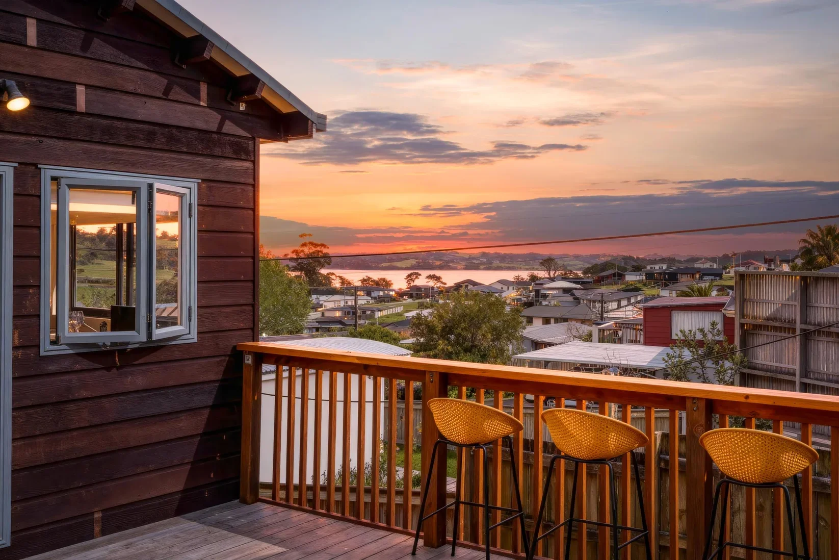 View from a wooden balcony at sunset, overlooking a neighborhood with houses and trees, with a body of water in the distance and colorful sky.