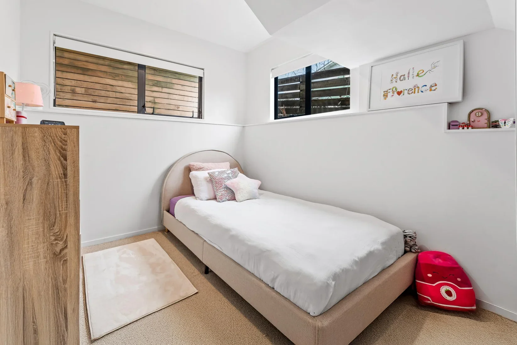 A small bedroom with white walls and beige carpet, featuring a single bed with a beige headboard, white bedding, pink and white pillows, and a plush star-shaped pillow. To the right of the bed is a red and pink plush backpack on the floor. There are 
