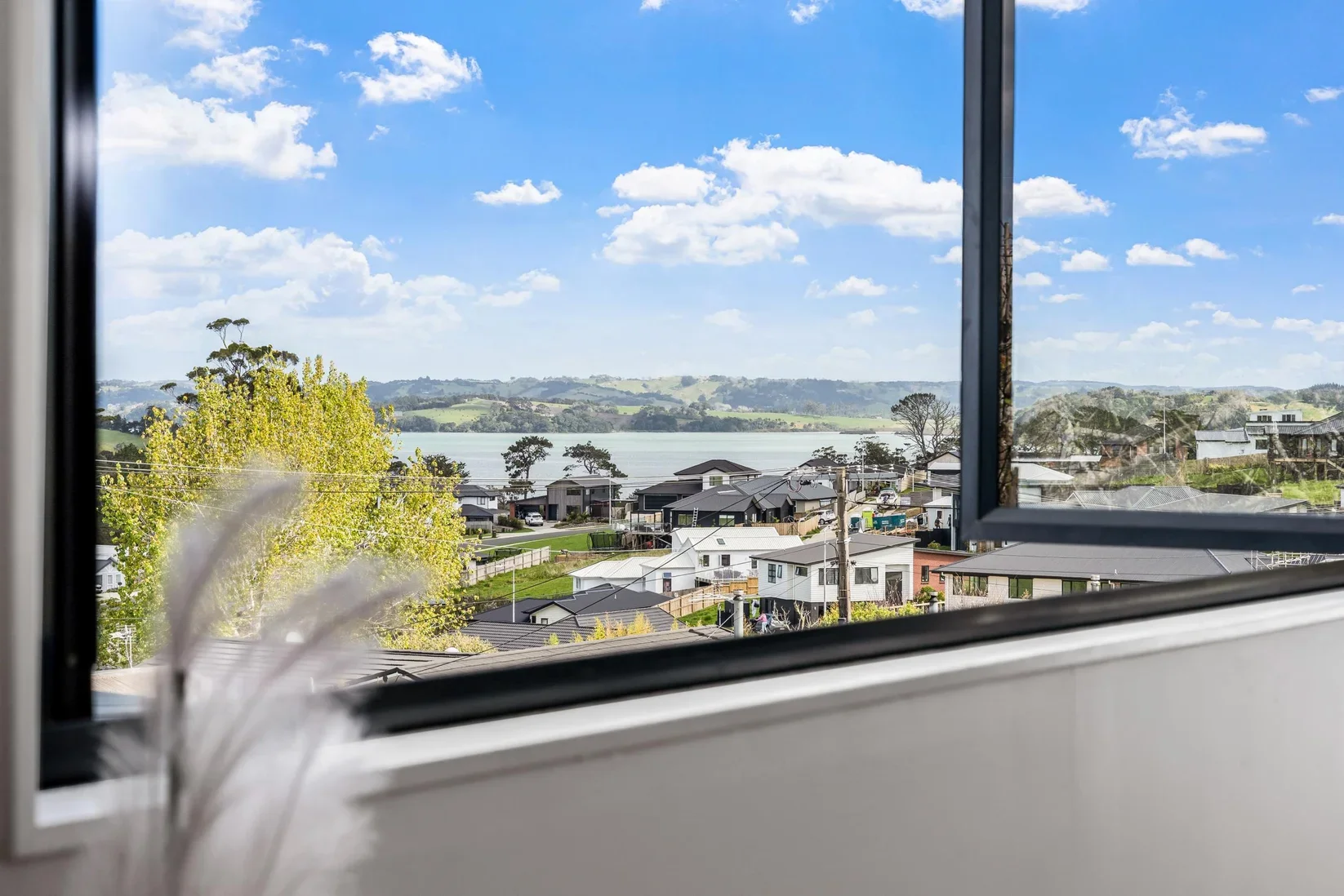 View of Snells Beach through an open window showing houses, trees, a body of water, and rolling hills under a partly cloudy sky.