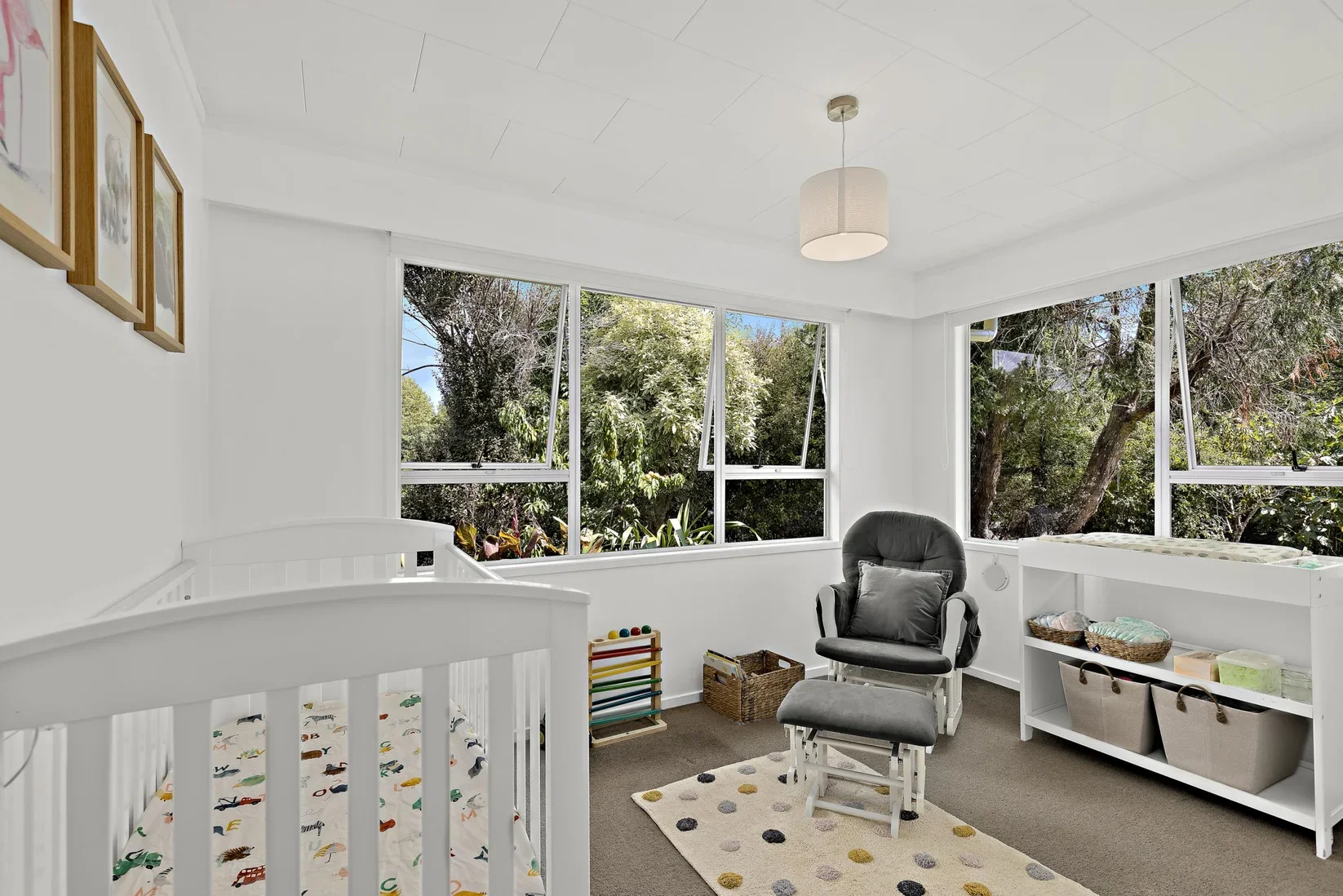 A nursery with white walls and lots of natural light from large windows, containing a white crib with colorful bedding, a black glider chair with a gray pillow, a white shelf with storage baskets, and a small colorful rug with polka dots.