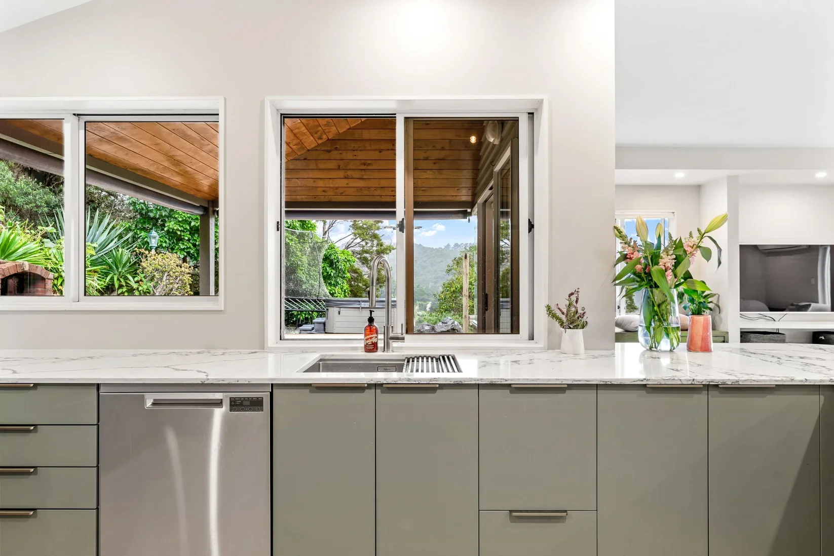 Modern kitchen with gray cabinets, marble countertop, stainless steel dishwasher, and a window showing lush green outdoor scenery with trees and plants.