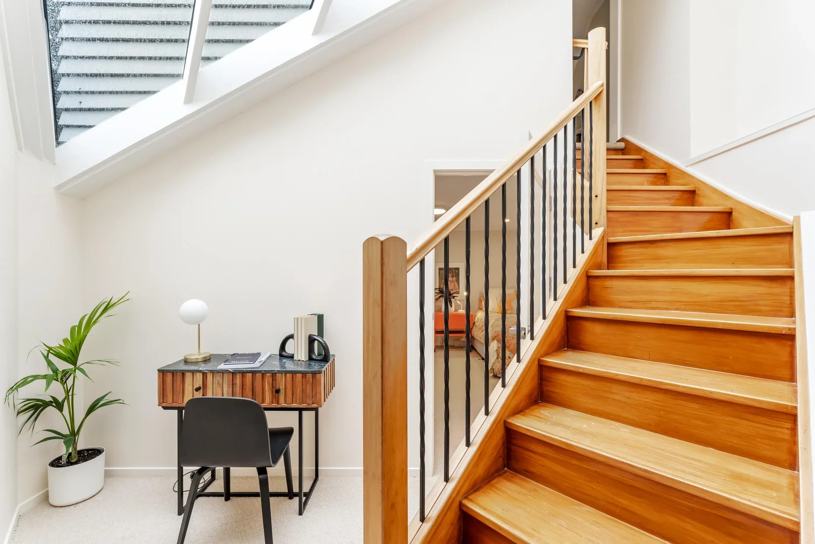 Interior of a home with a wooden staircase, small desk with a black chair, potted plant, and decorative lamp.