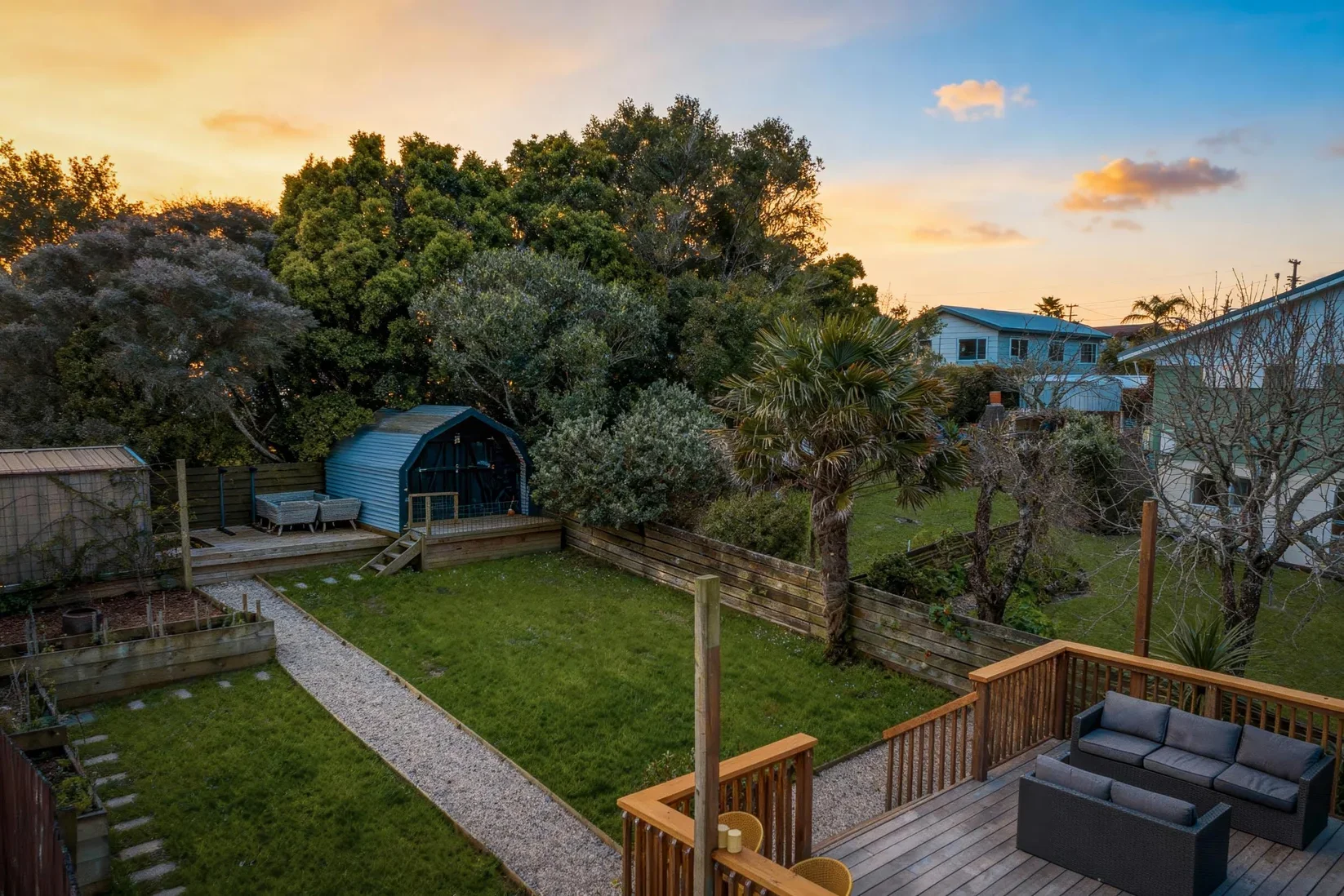 Residential backyard during sunset with green lawn, gravel pathway, outdoor seating, trees, and houses in the background.