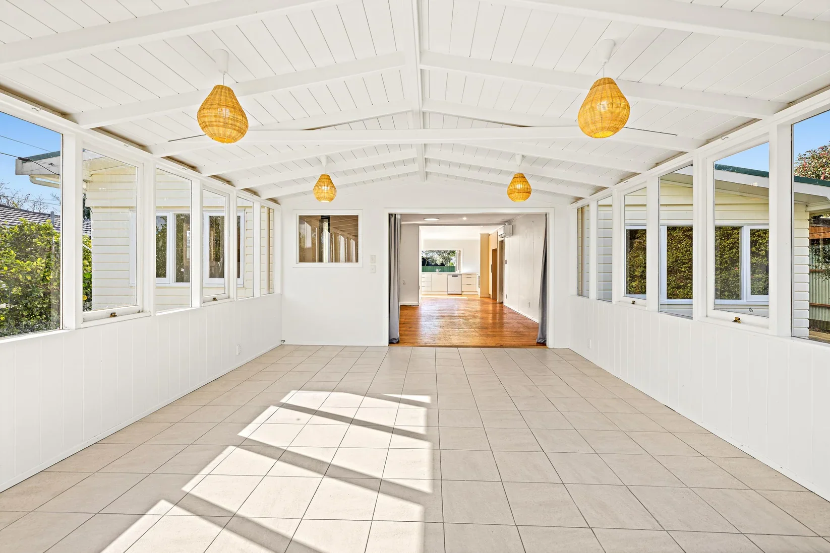 Bright enclosed porch with large windows, white walls and ceiling, beige tiled floor, and hanging wicker light fixtures, leading into a hardwood-floored living area with an open kitchen in the background.