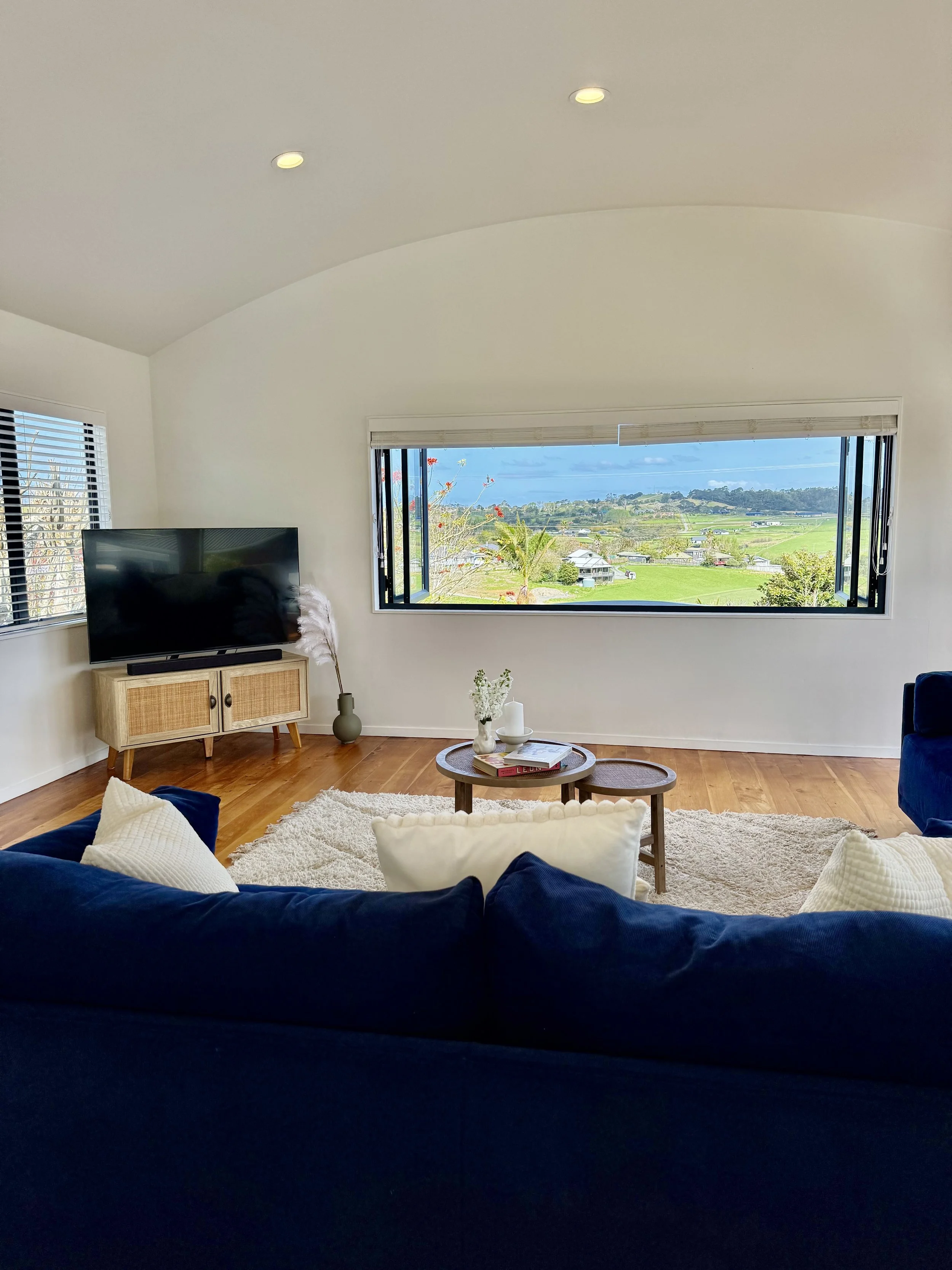 Living room with large window showcasing a scenic outdoor view, dark blue sofa with white pillows, coffee tables with decor, wooden TV stand with television, beige rug, and light-colored walls.