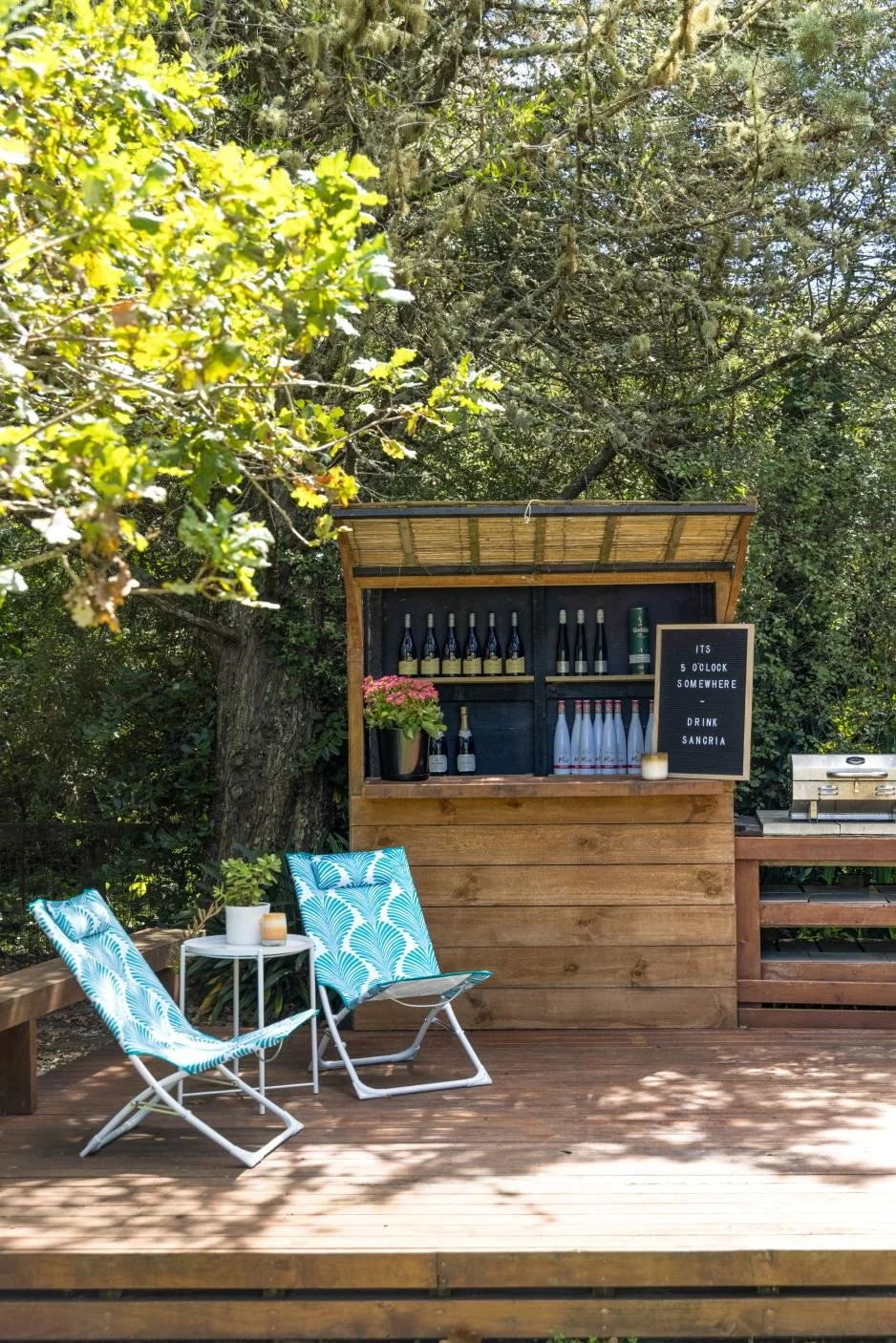 Outdoor bar setup with two blue patterned chairs, a small white table with potted plant, bottles of alcohol on a wooden bar, a blackboard sign, and a grill in a green, shaded backyard.