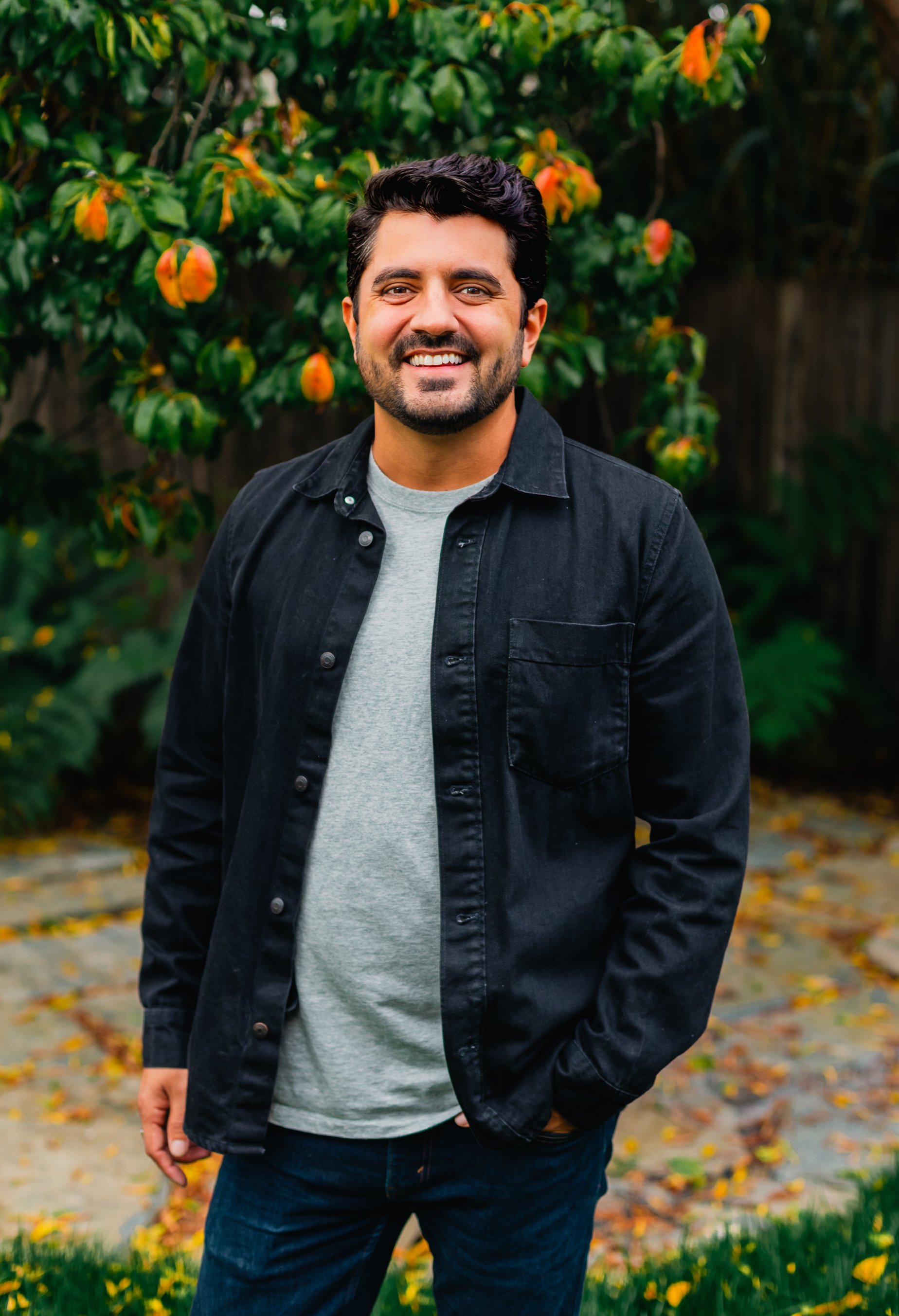 Smiling man with dark wavy hair and a short beard wearing a black button-up shirt over a gray t-shirt, standing outdoors in front of green foliage and a backyard garden pathway.