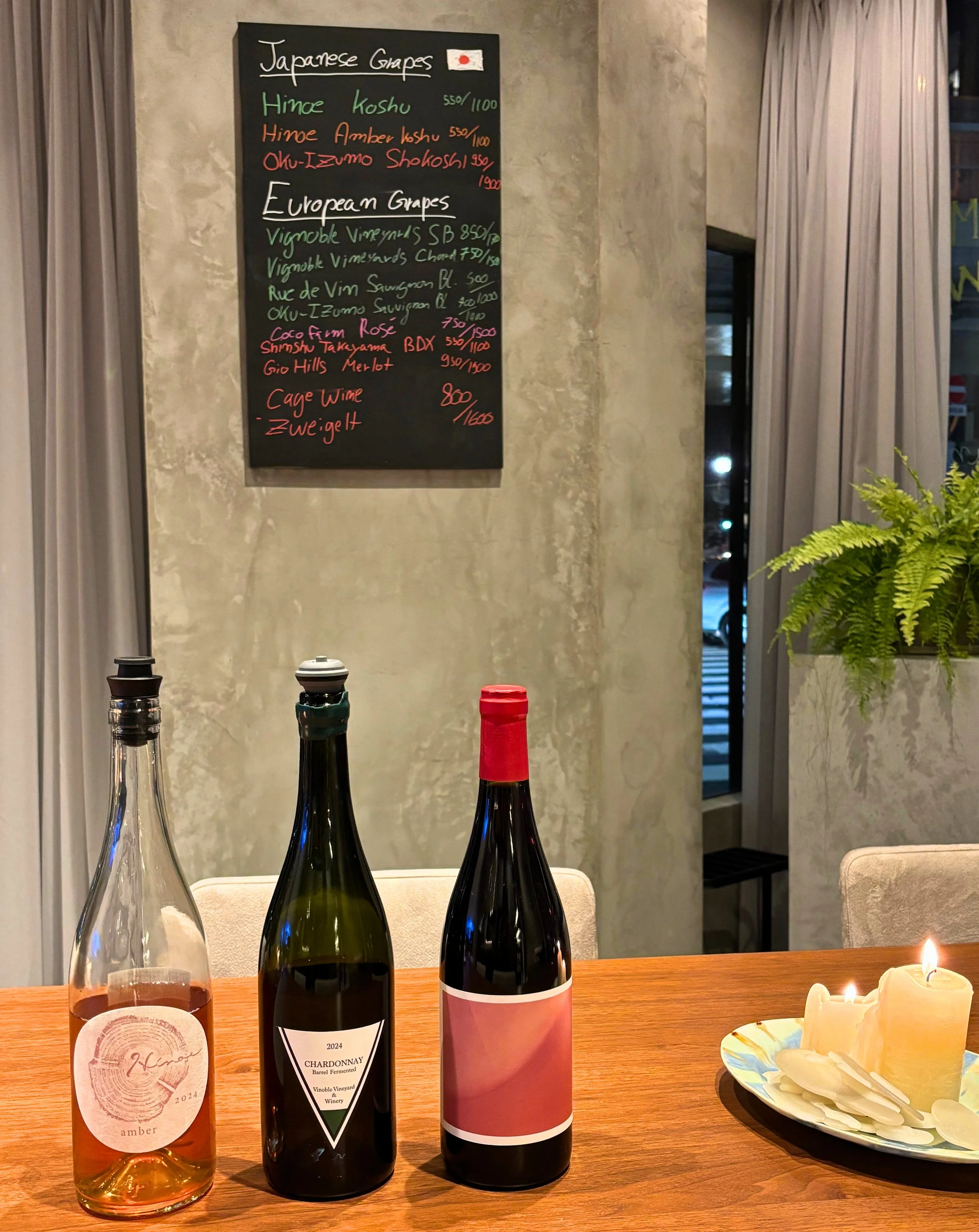 Opened Japanese wine bottles with daily by-the-glass selections on the blackboard at the THREE GRAPES wine bar in Tokyo.