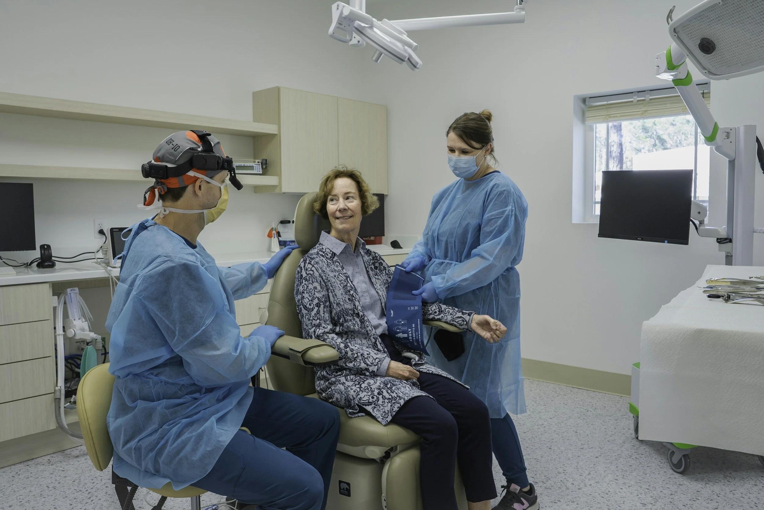 A female doctor and two medical professionals are attending an elderly woman patient in a doctor's office or clinic. The patient is sitting on a medical chair, smiling, while the healthcare workers are standing nearby, wearing blue protective gowns and masks. One of the professionals is wearing a headlamp and appears to be preparing for or performing a procedure.