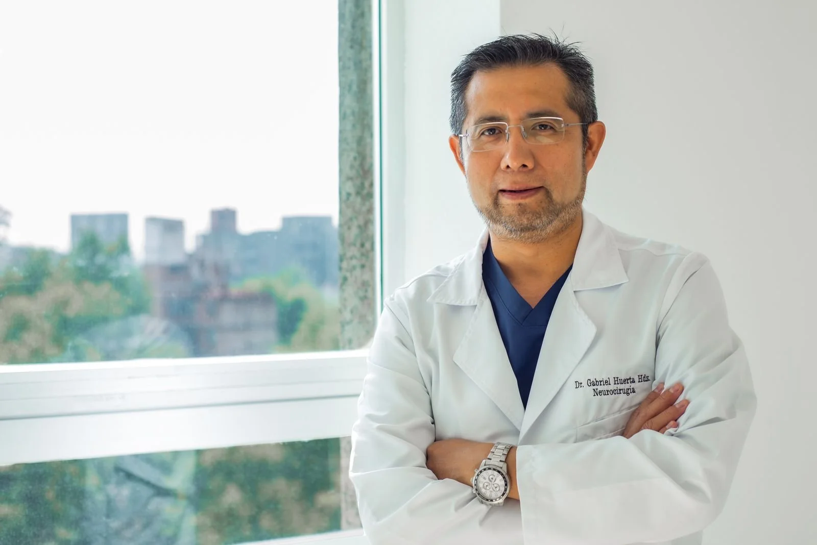 A male doctor wearing glasses and a white coat with a name tag, standing with arms crossed in a well-lit room with a window showing trees and buildings outside.