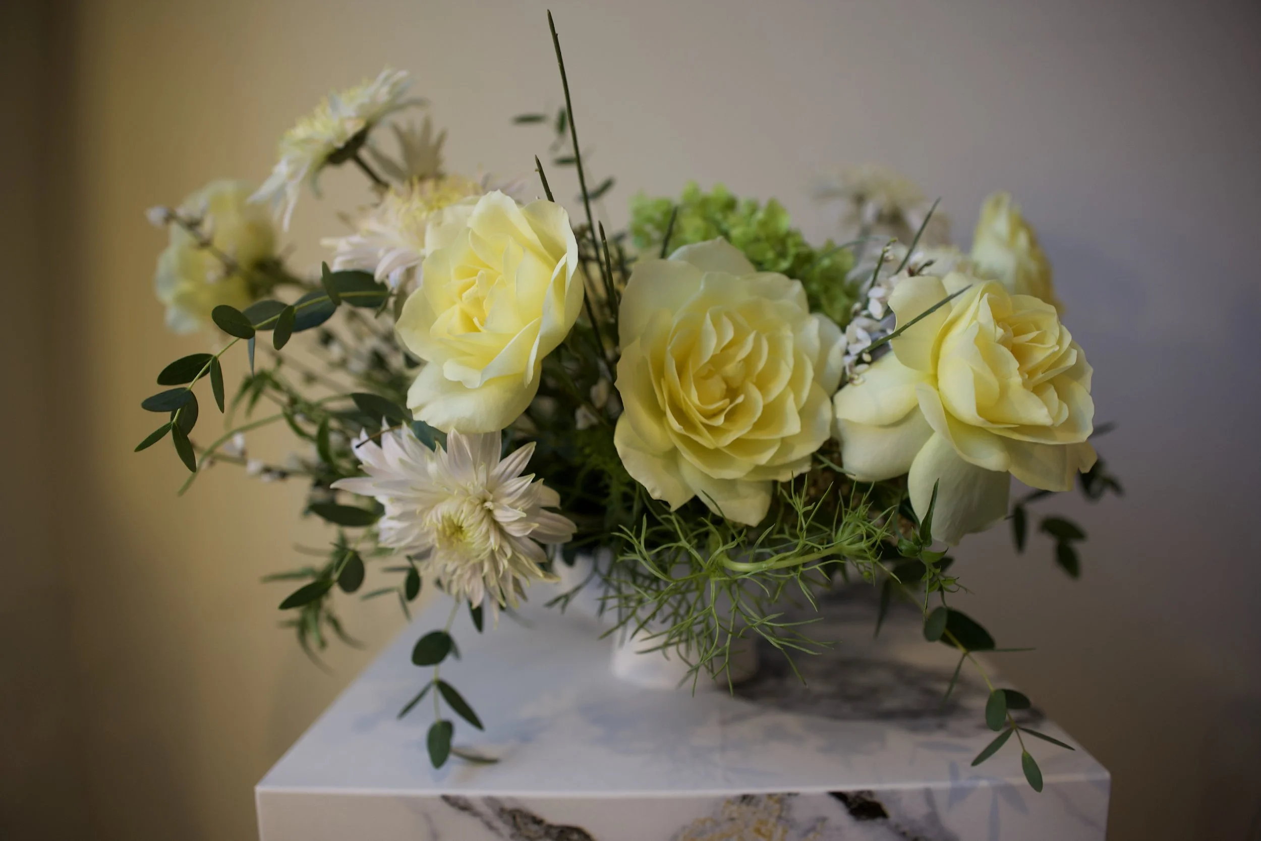 A floral arrangement with yellow roses, white daisies, and greenery on a marble surface. Perfect arrangement for weddings as a low centerpiece for your long square tables