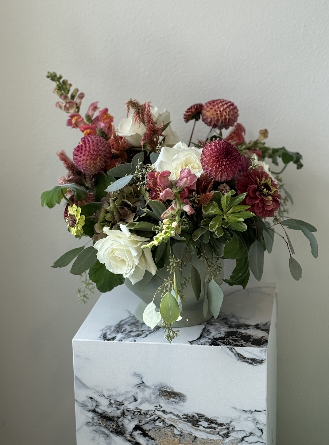 A floral arrangement with white roses, pink and red dahlias, and various greenery in a gray vase on a marble pedestal against a plain wall.