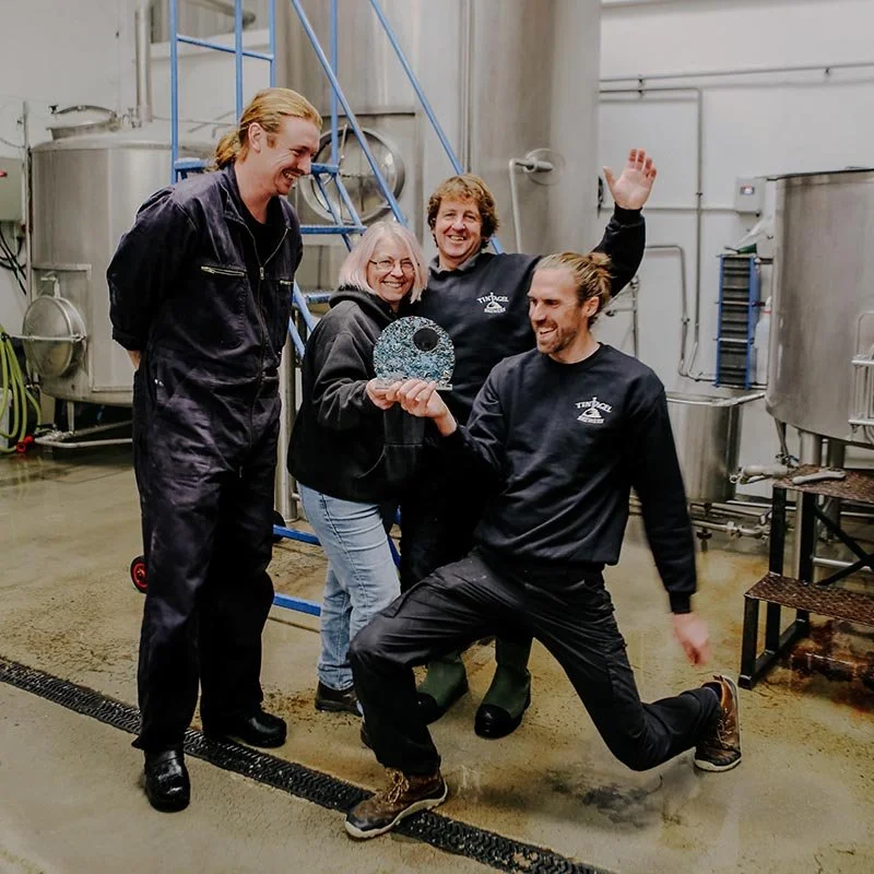 The Tintagel Brewery team celebrates winning an award. Four smiling staff members—two men in dark workwear, one woman holding the glass award, and another man in a celebratory pose—stand together amongst stainless steel brewing equipment.