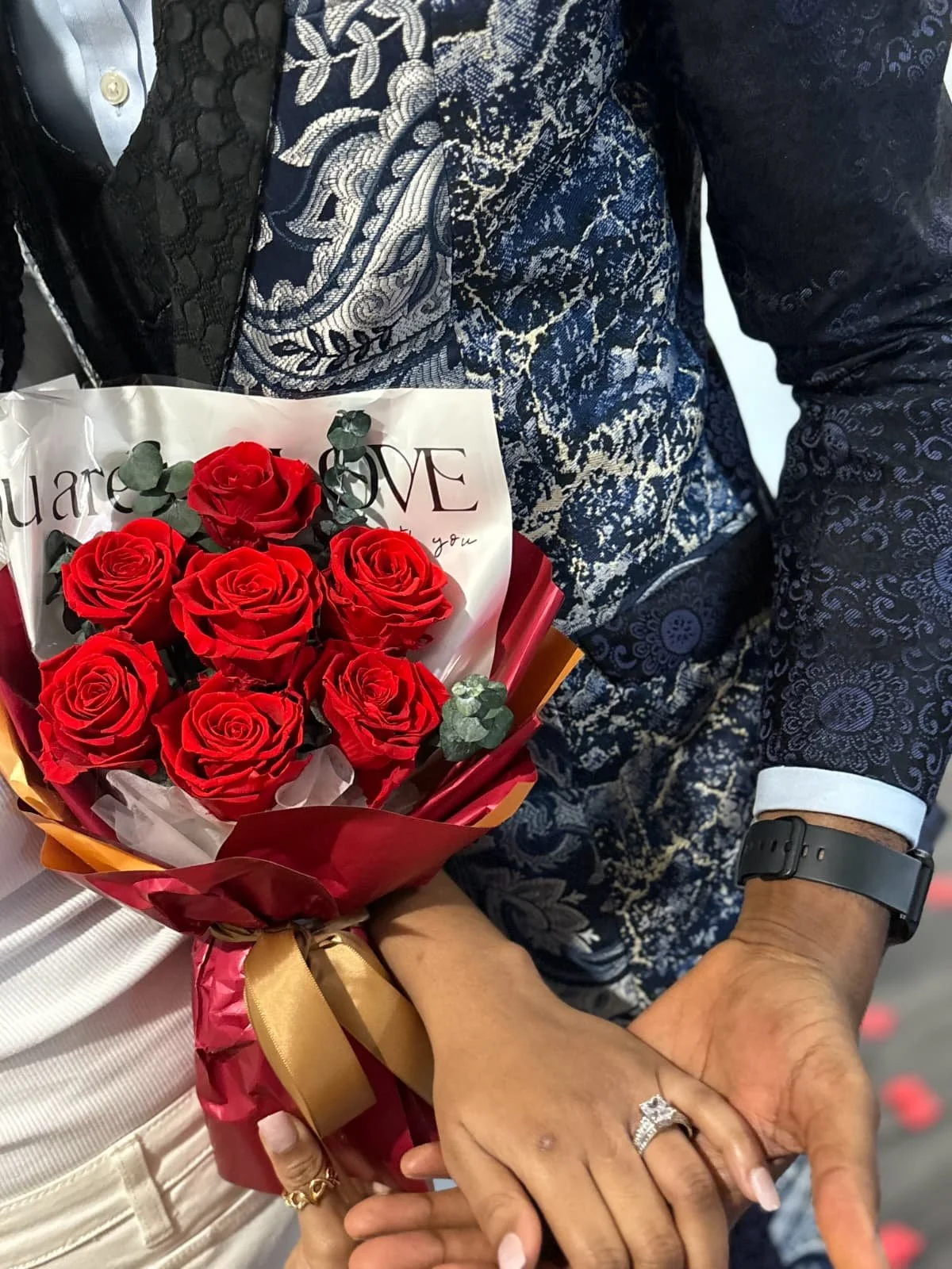 A person holding a bouquet of red roses with a 'love' message, accompanied by a woman with a wedding ring on her finger, both holding hands.