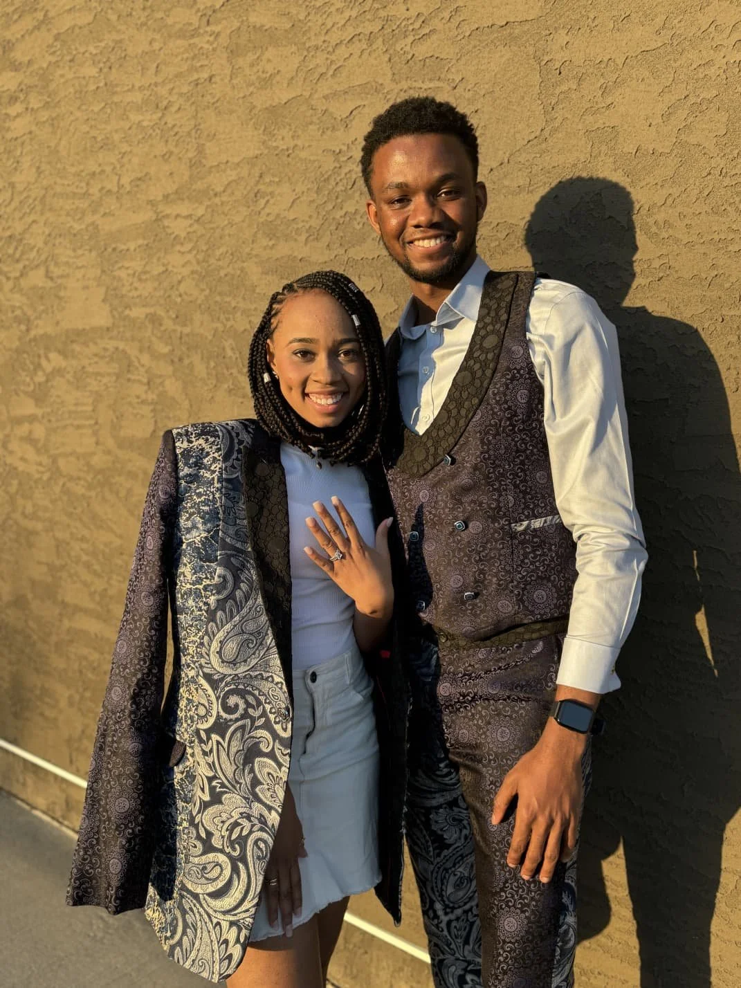 A smiling couple standing outside against a textured beige wall, the woman showing her wedding ring, the man wearing a light-colored shirt and vest, both dressed stylishly.