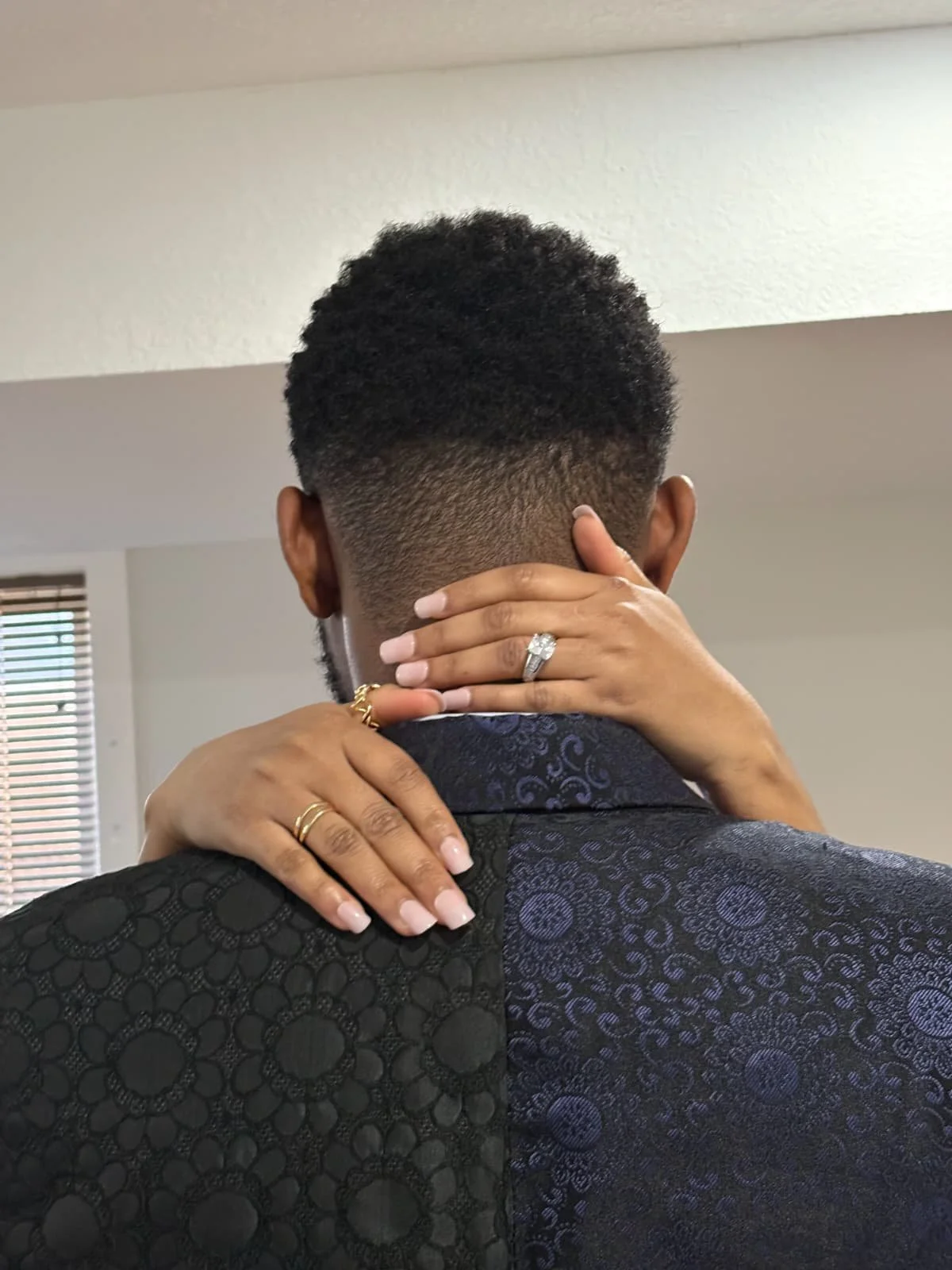 A couple embraces, showing the woman's hands with a ring and wedding bands resting on the man's neck and shoulder.