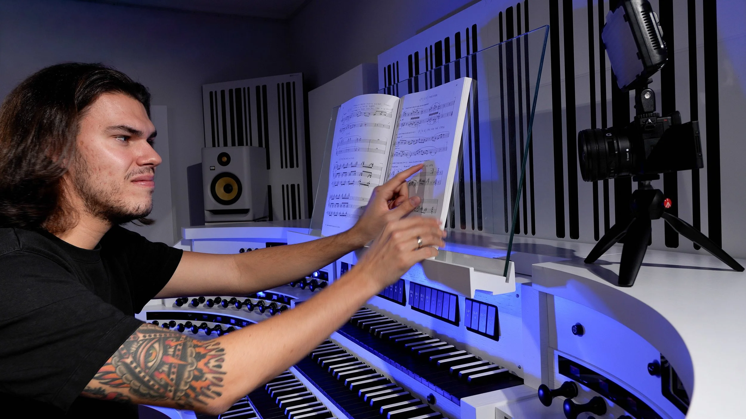 Giulio Lucciola explaining organ music at a modern digital organ console during an educational recording, with sheet music and camera visible in the studio.